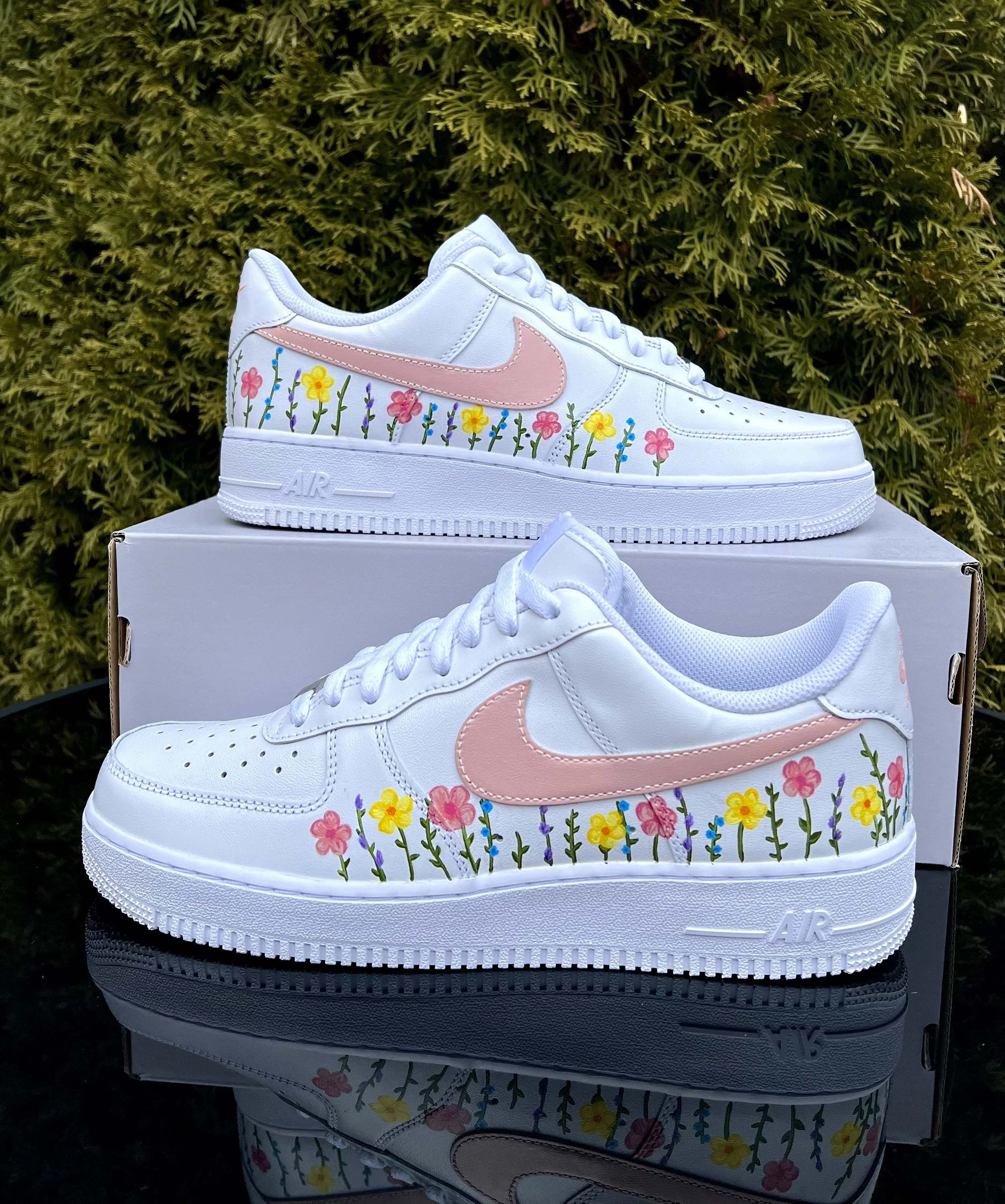 White Nike Air Force 1 sneakers with pink swoosh and colorful floral designs on the sides, placed on top of a white box outdoors with greenery in the background.