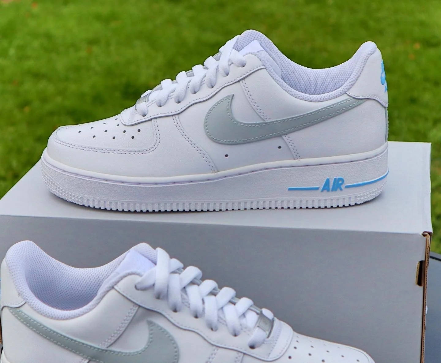 A white Nike Air Force 1 sneaker with a gray Nike swoosh logo on the side, placed on a white box with a blurred grassy background.