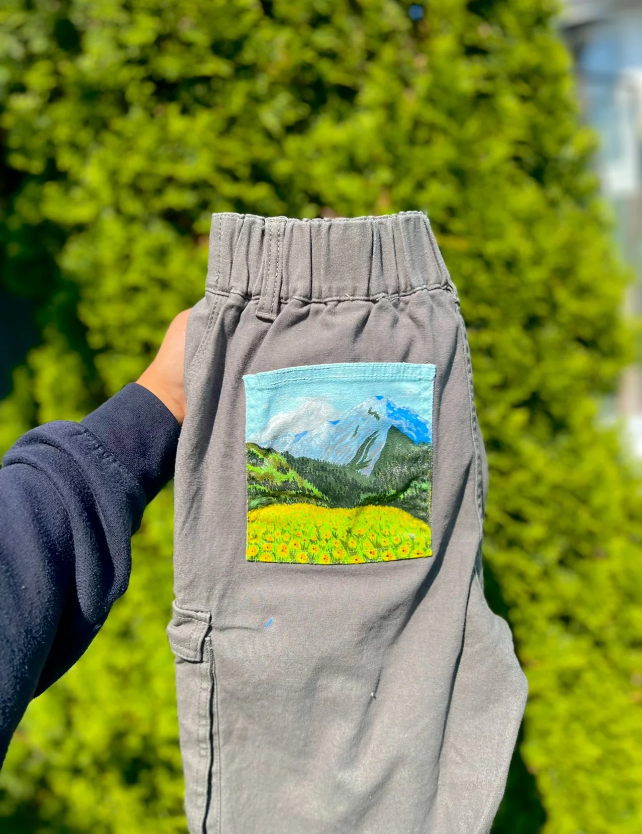 Gray pants with a colorful mountain landscape patch embroidered on the back pocket, being held up outdoors with green foliage in the background.
