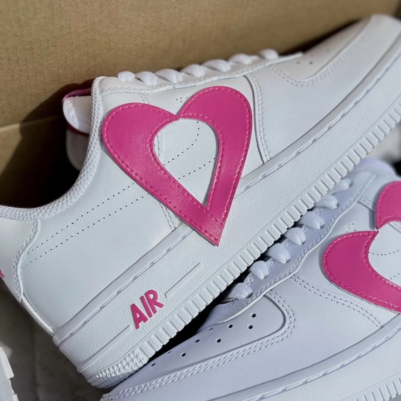 Close-up of a white sneaker with pink heart-shaped accents on the side and the word 'AIR' printed in pink near the sole.