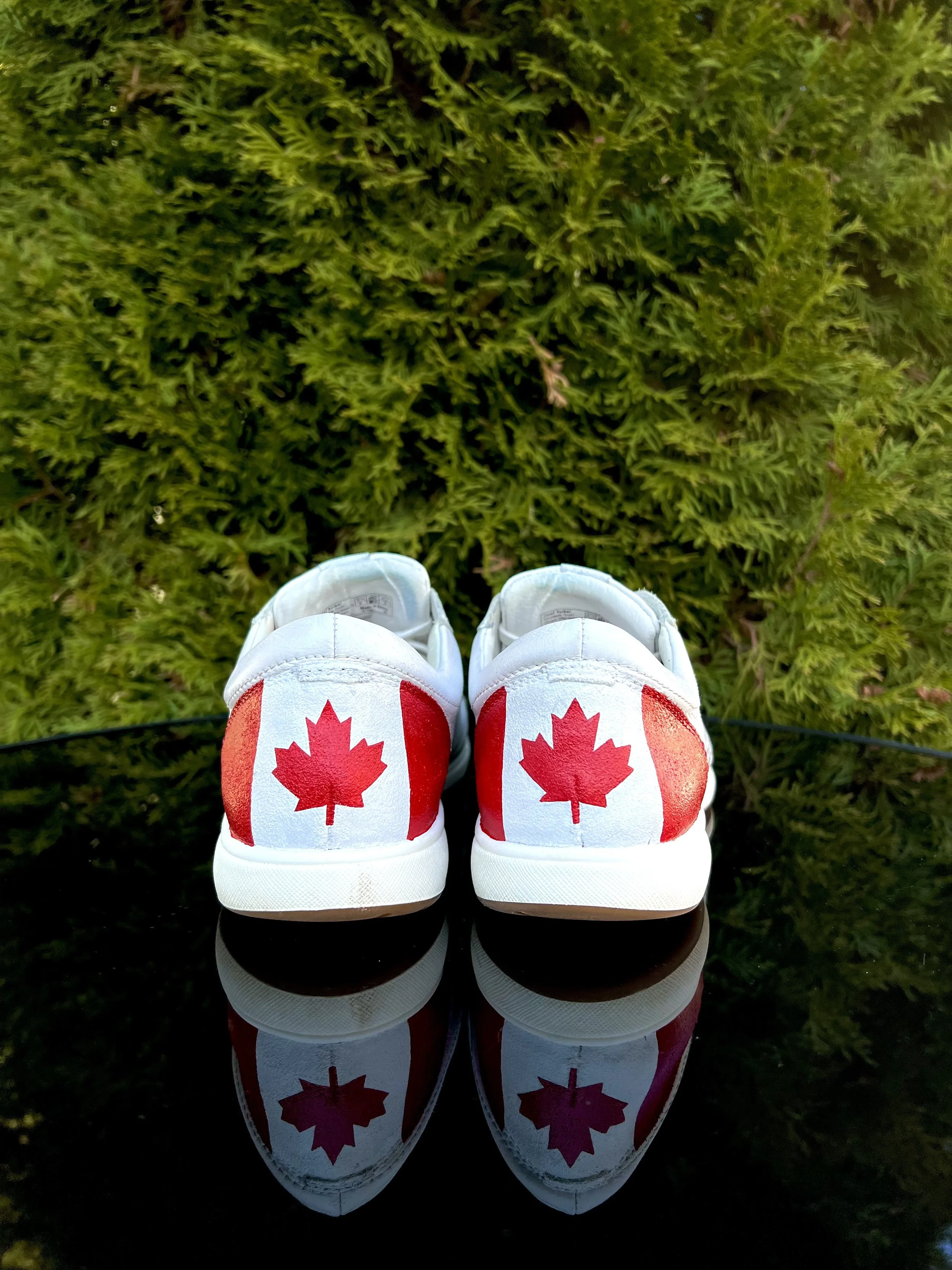 A pair of white sneakers with red Canadian maple leaf designs on the back, reflected on a shiny black surface, with green foliage in the background.