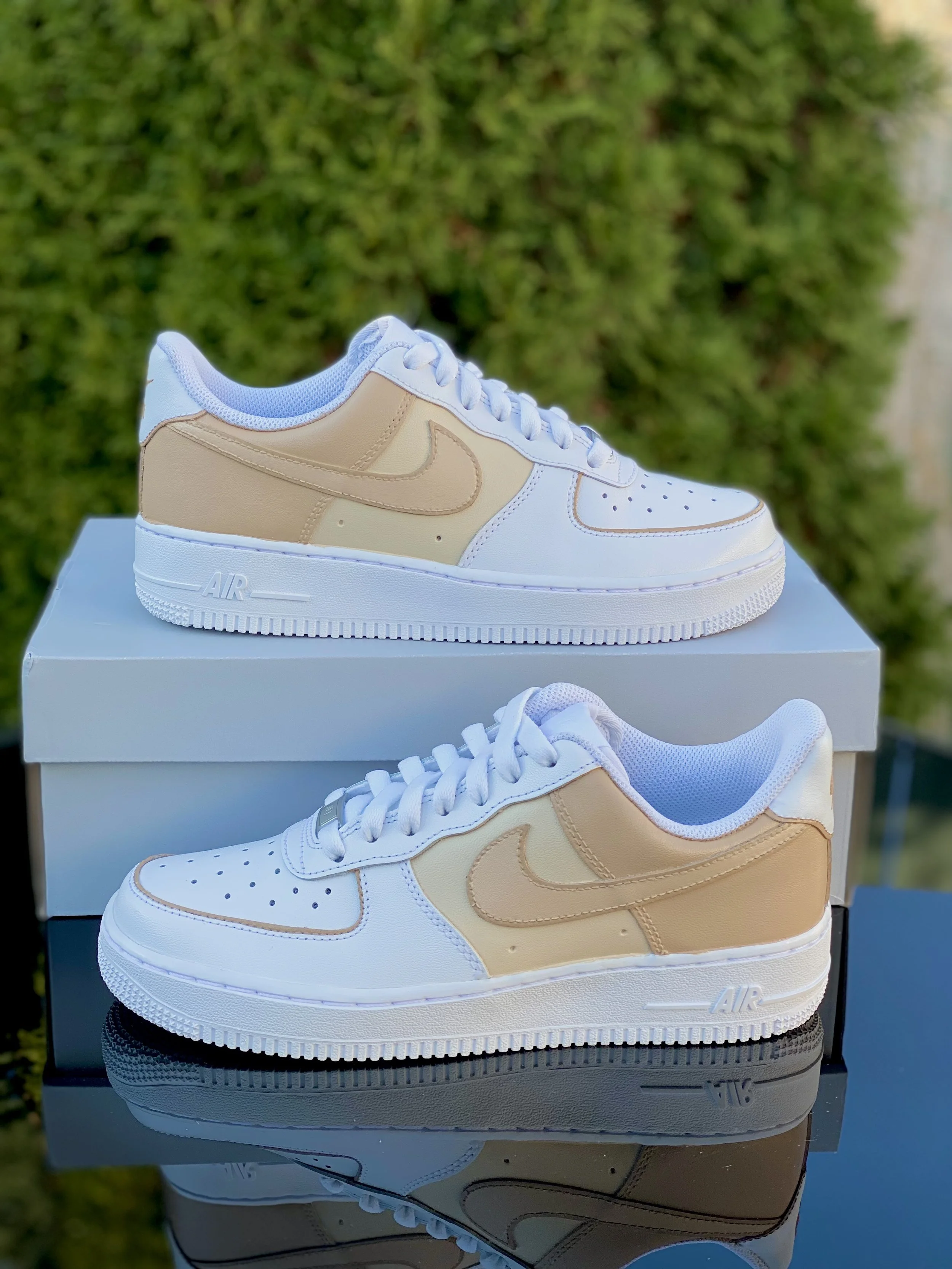 Beige and white Nike Air Force 1 sneakers on white shoe box with greenery in the background.