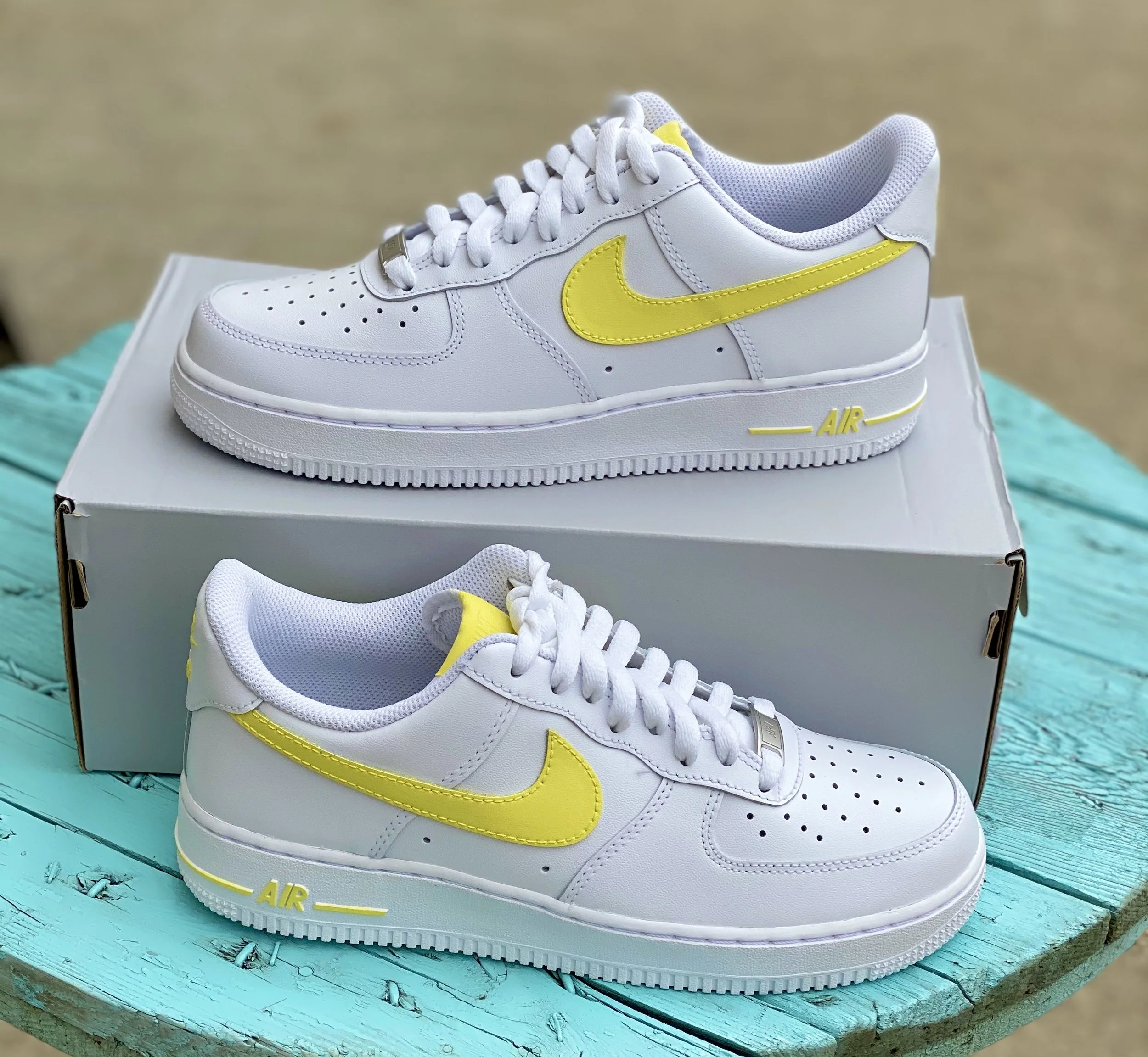 White Nike Air Force 1 sneakers with yellow accents, placed on a white box, on a teal wooden surface.