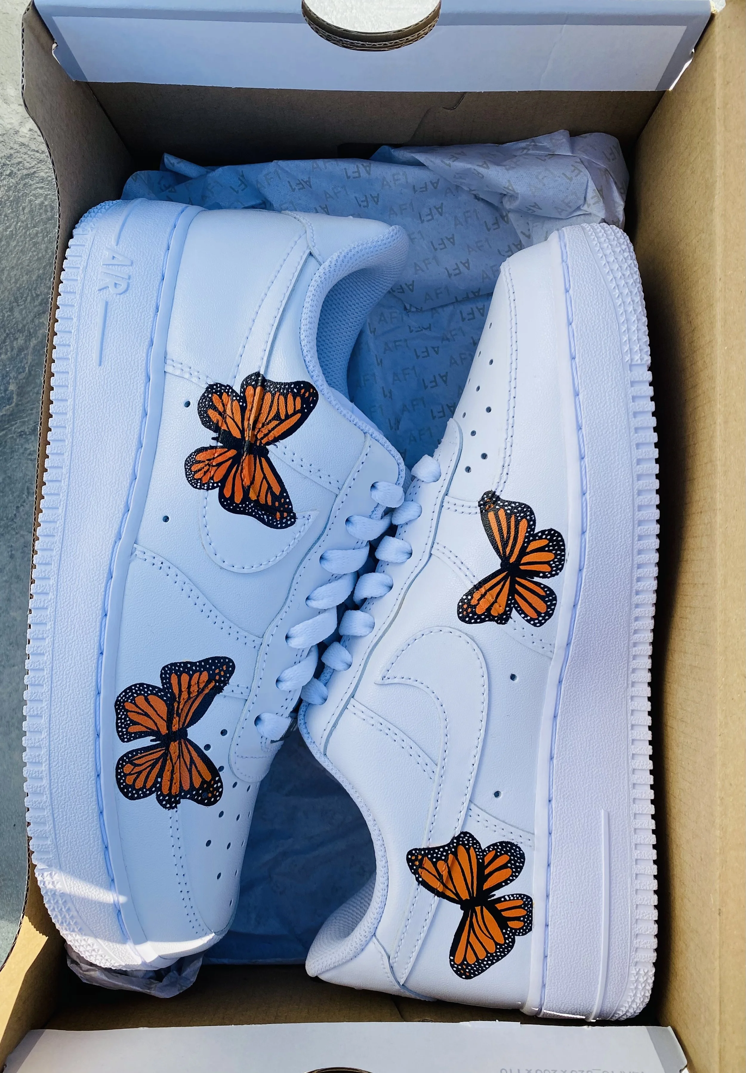 White sneakers with orange and black butterfly designs on the side, inside a brown shoebox with tissue paper.