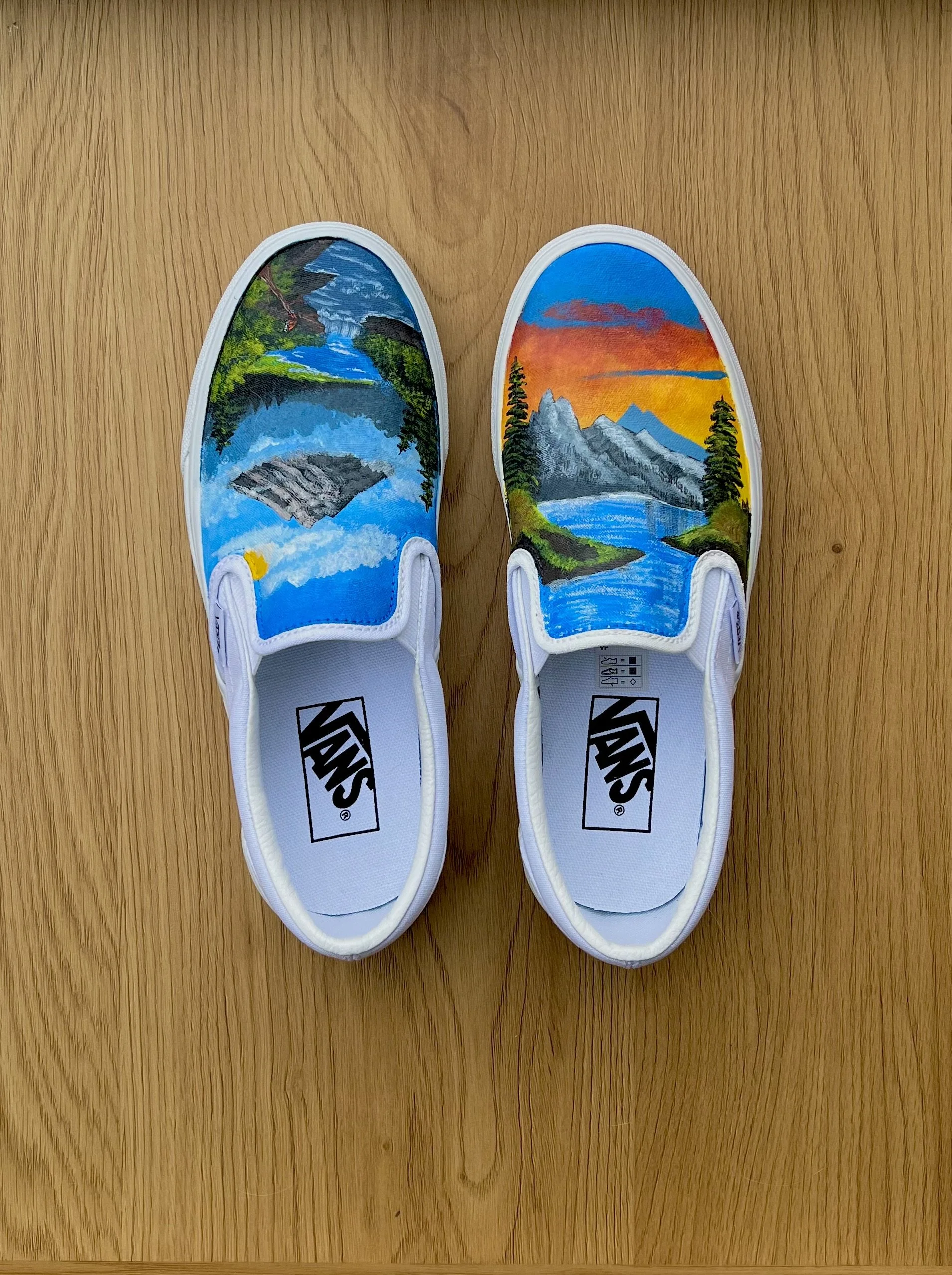 Pair of white slip-on sneakers with hand-painted scenic landscape designs featuring mountains, rivers, trees, and sunset.