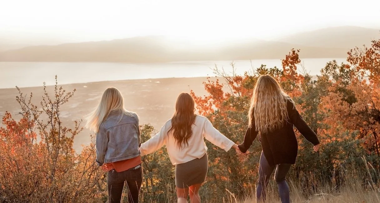Three women walking hand in hand through an autumn field at sunset, supporting each other.