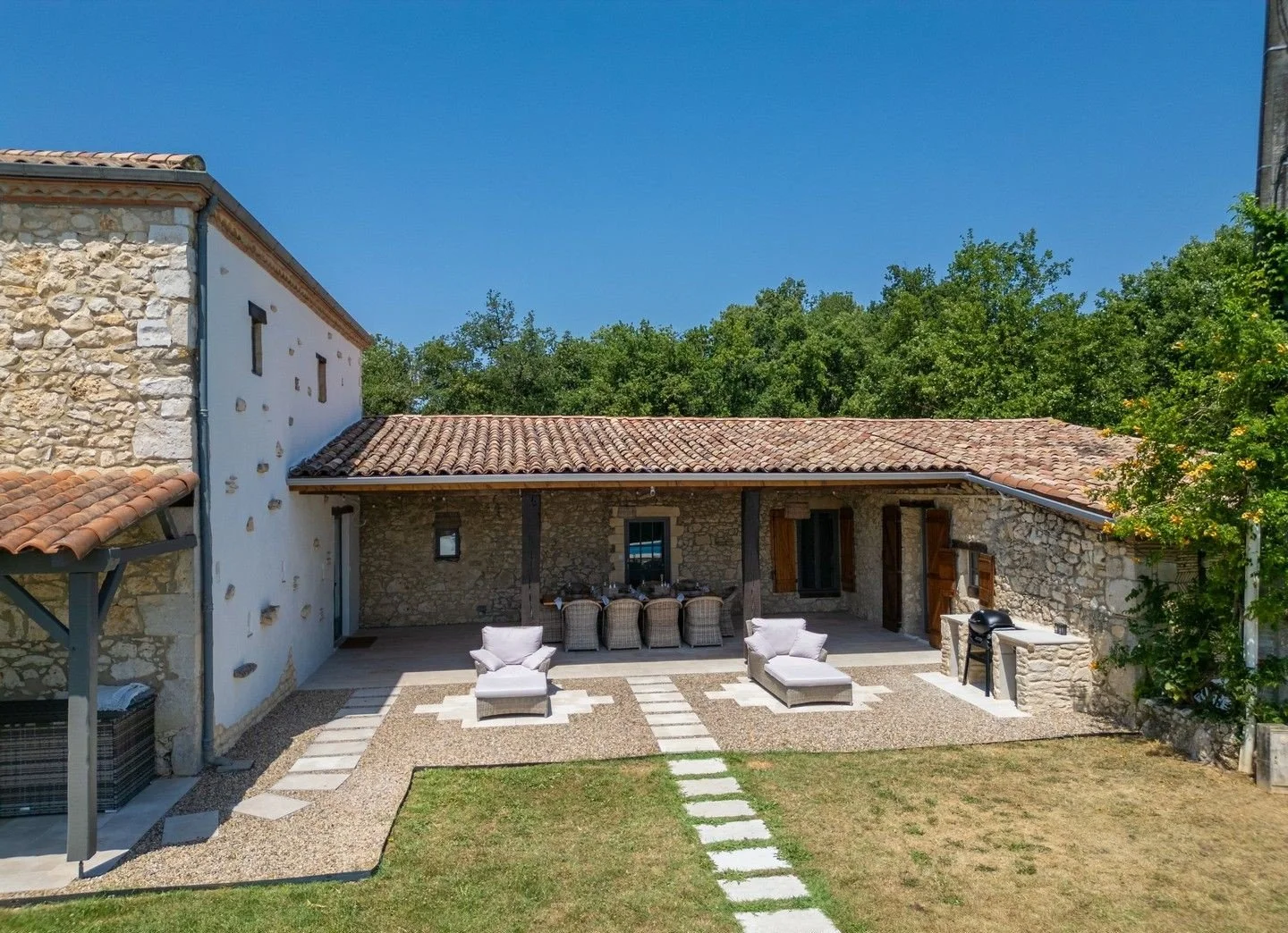 Stone house with red tile roof and outdoor patio area with lounge chairs, dining table, and barbecue grill, surrounded by greenery under a clear blue sky.