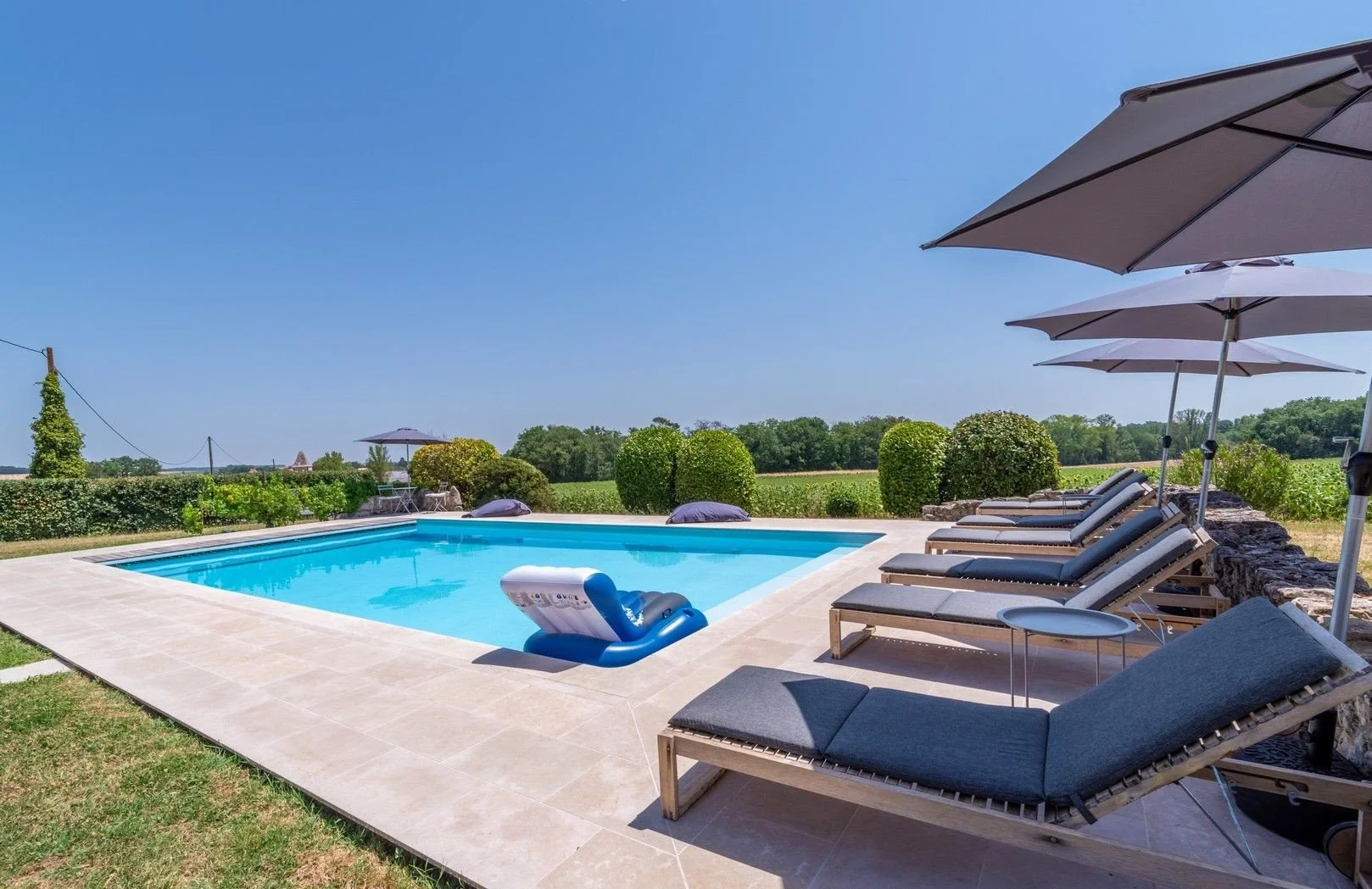 An outdoor swimming pool surrounded by lounge chairs and umbrellas with a view of green fields and trees under a clear blue sky.
