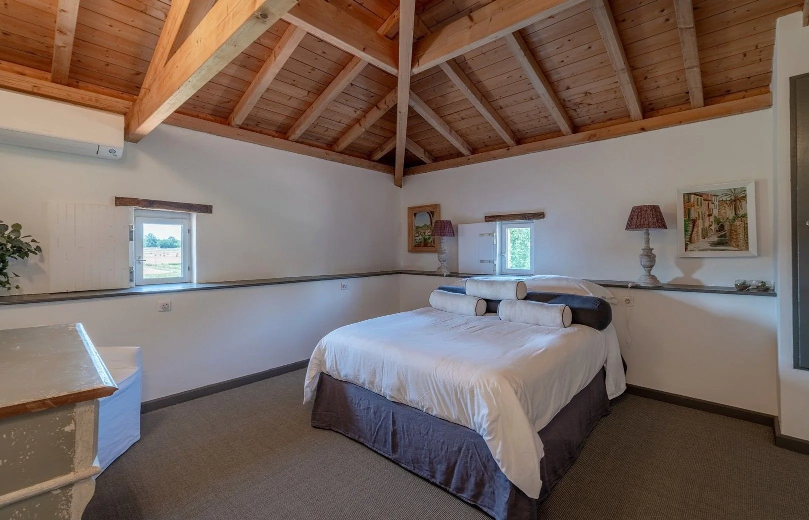 A cozy bedroom with a sloped wooden ceiling and small windows, a bed with white linens and rolled pillows, bedside lamps, and framed artwork on white walls.