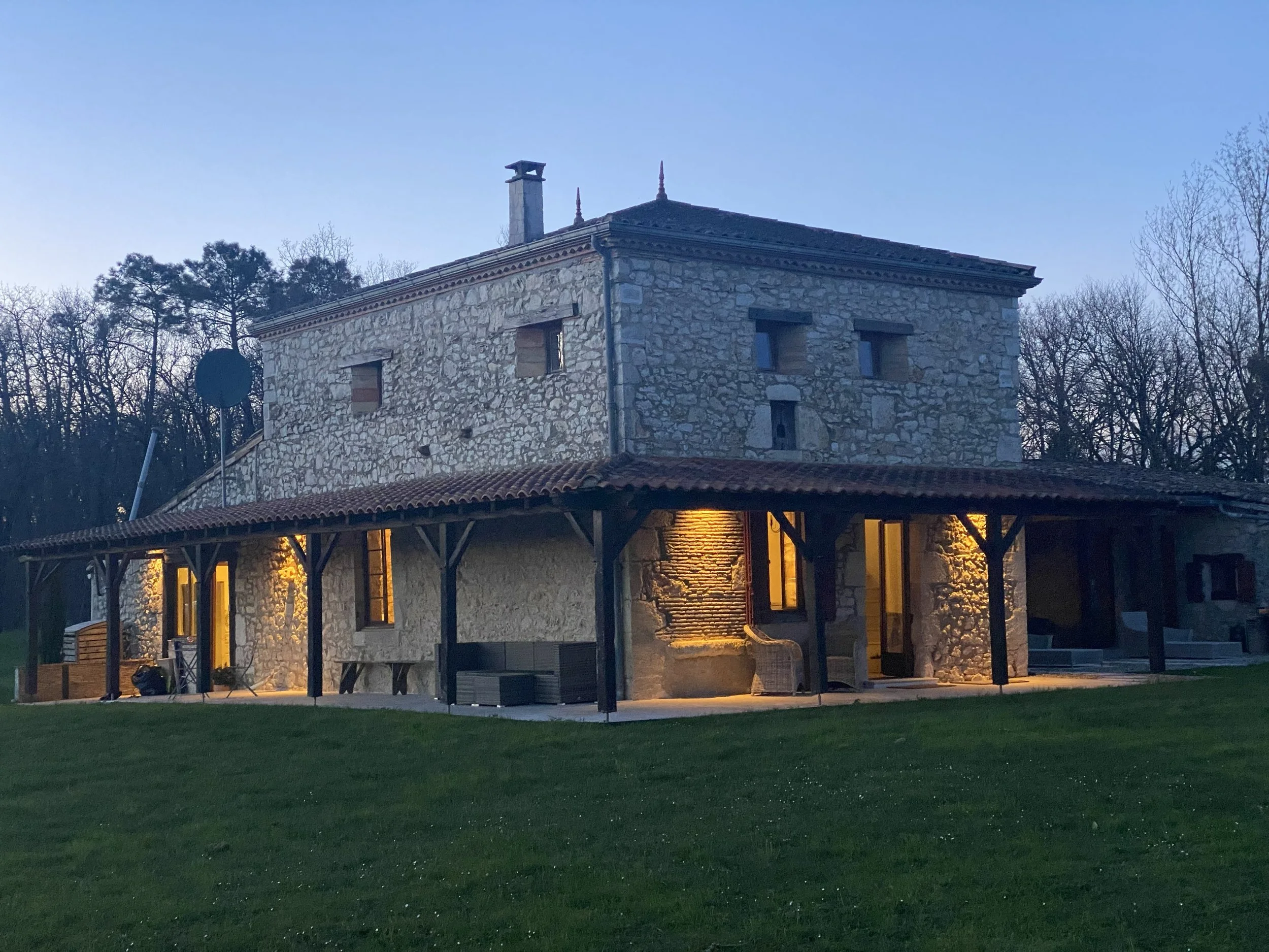 A stone house with a sloped roof, illuminated from inside, surrounded by a wooden porch with columns, set against a backdrop of trees during dusk.