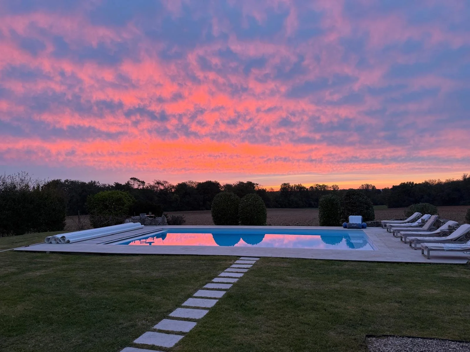 A backyard swimming pool surrounded by lounge chairs, with a sunset sky filled with pink and purple clouds.