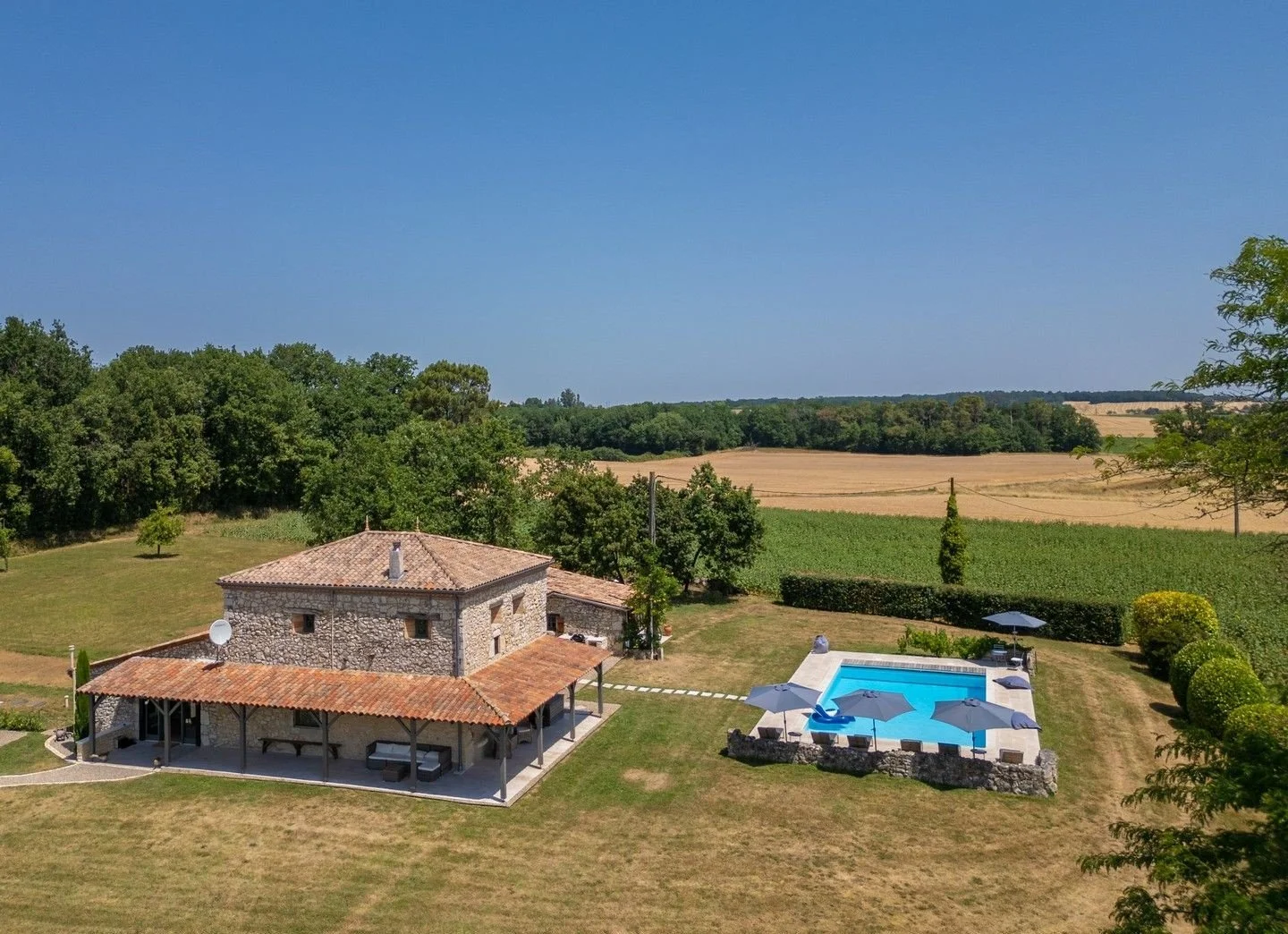 A countryside property with a stone house, a swimming pool with umbrellas, and a large grassy yard, surrounded by fields and trees under a clear blue sky.