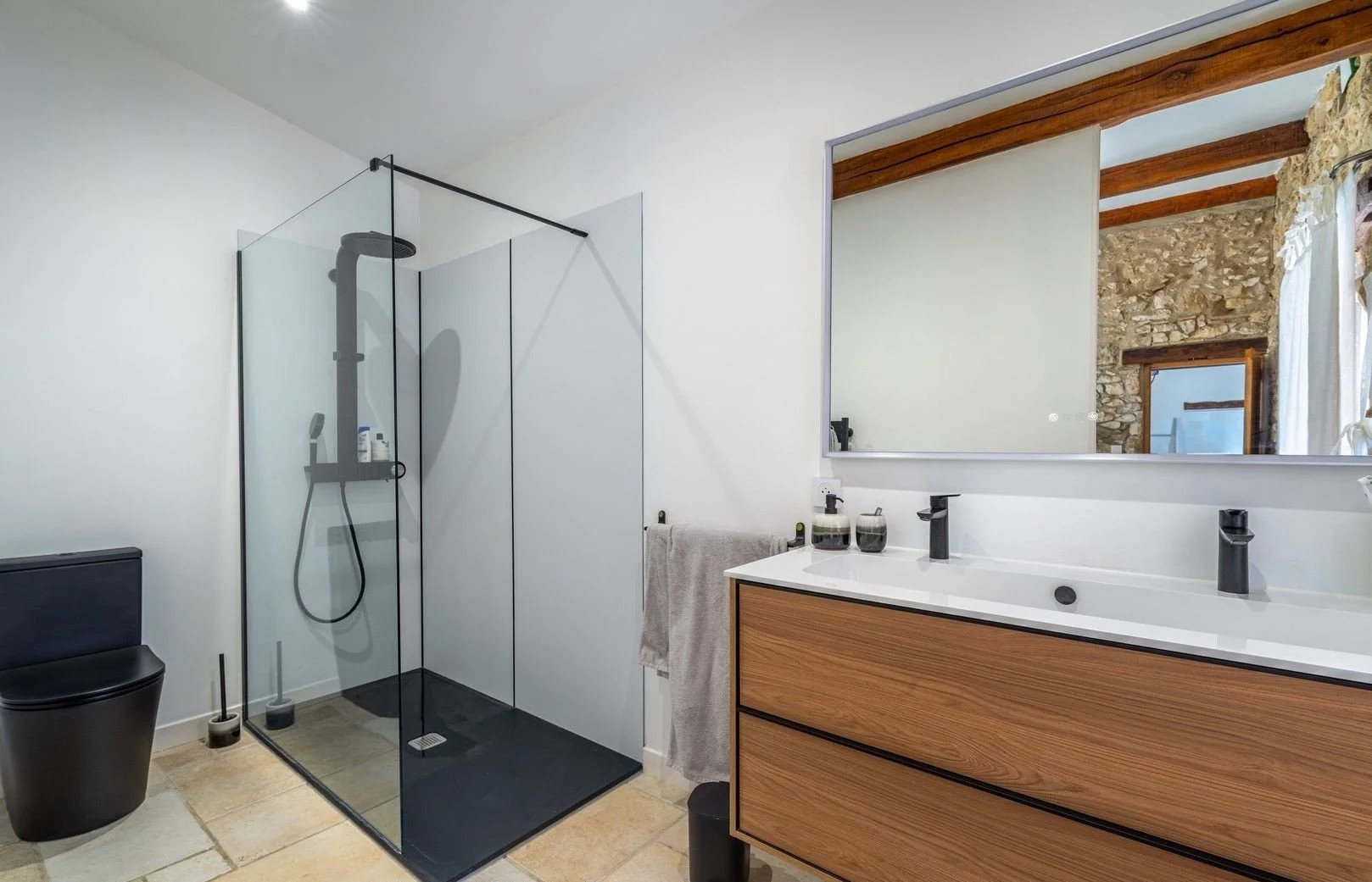 Modern bathroom with a walk-in shower with a black drain and toiletries on a shelf, a black toilet, a wooden vanity with a white countertop, a large mirror, and a towel hanging on the wall.