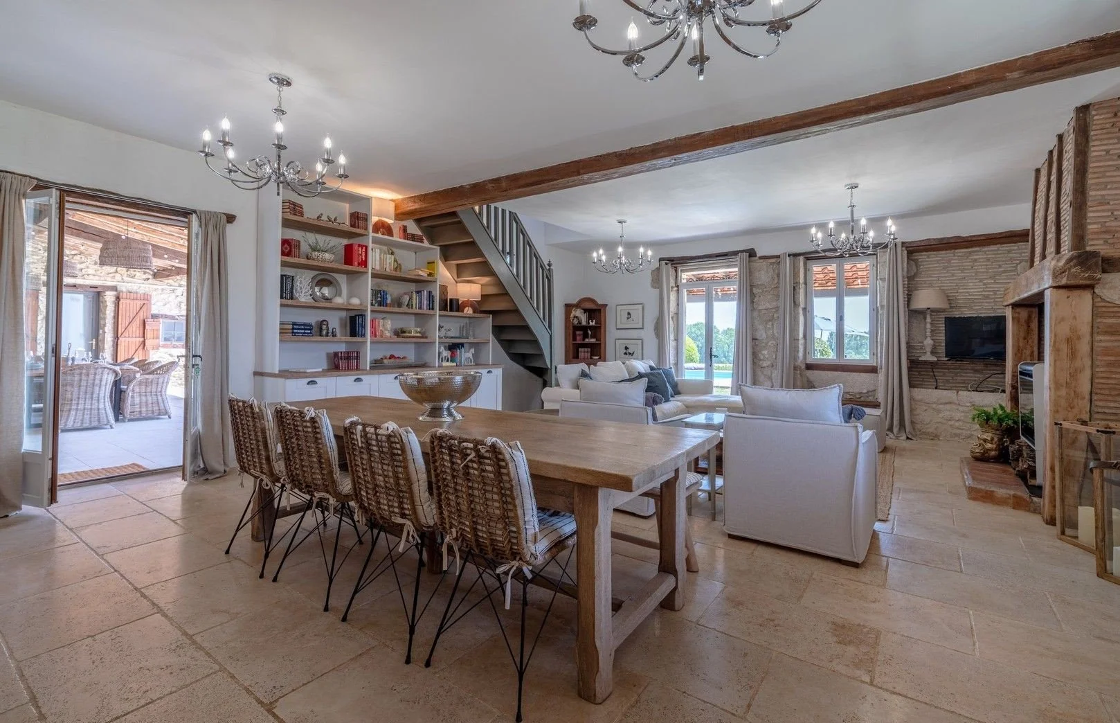 Spacious living and dining area in a rustic style home with tile flooring, a wooden dining table with woven chairs, white sofas, a stone and wood fireplace, and ample natural light from large windows and sliding glass door leading to an outdoor patio.