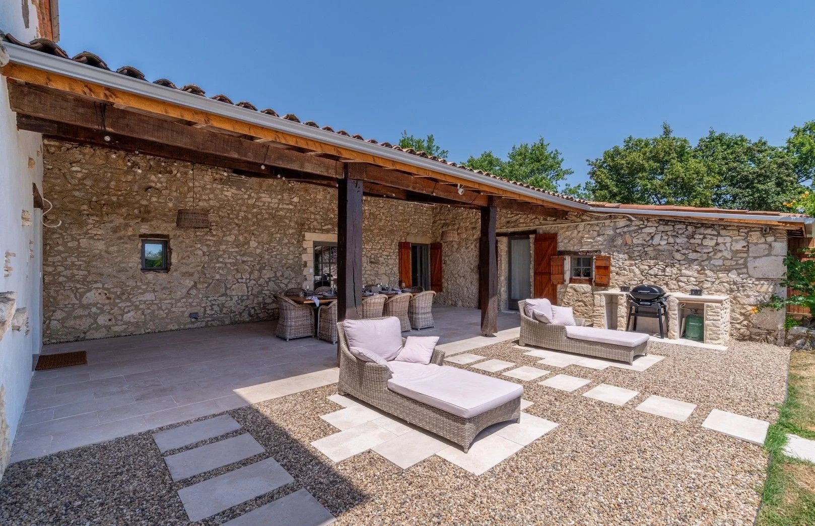 Outdoor patio area with wicker lounge chairs, a dining table with chairs, a stone barbecue grill, and a stone wall with wooden shutters, under a wooden roof.