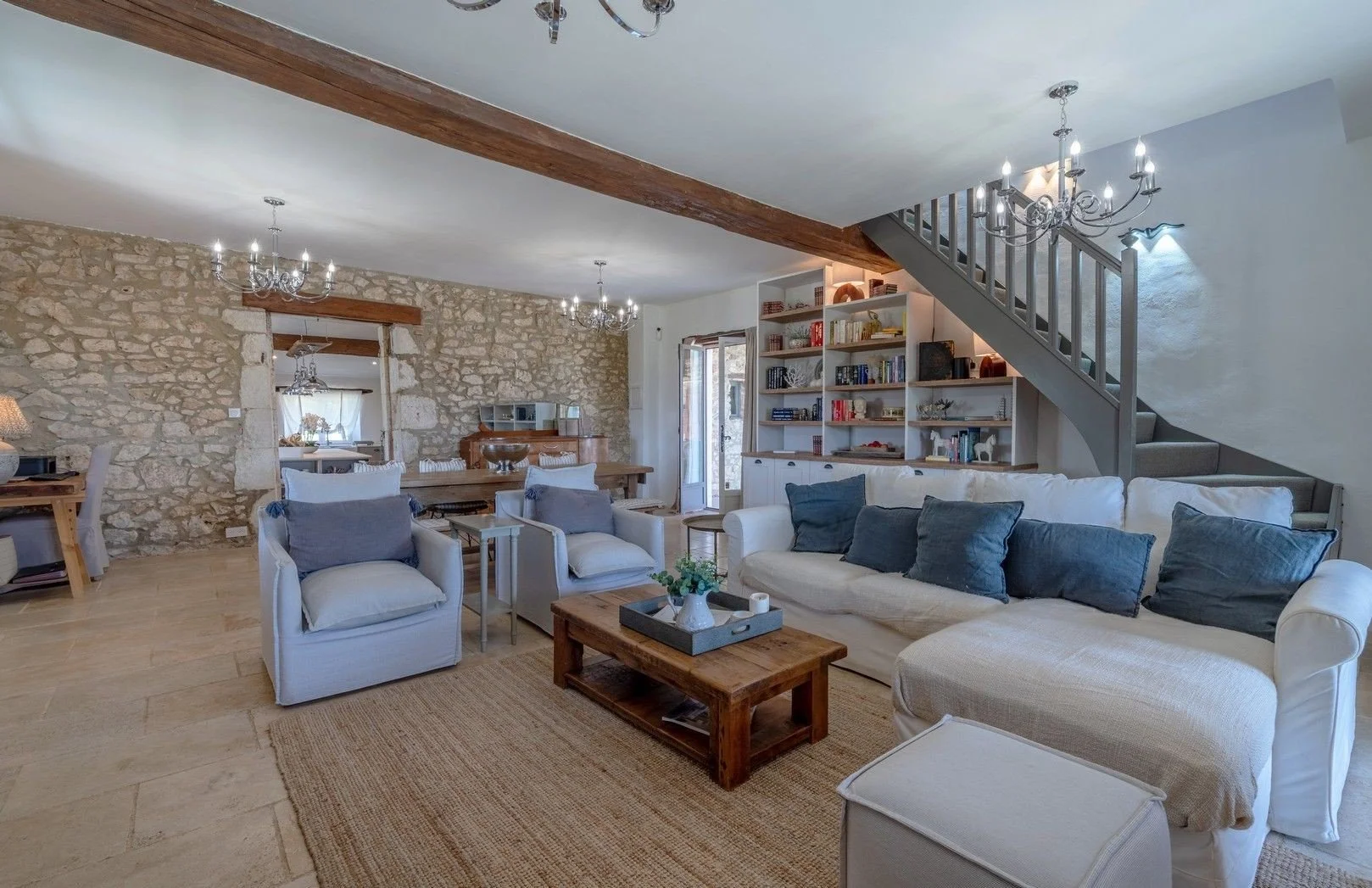 Living room with light-colored furniture, blue cushions, stone wall, white built-in bookshelves, staircase, and chandeliers.