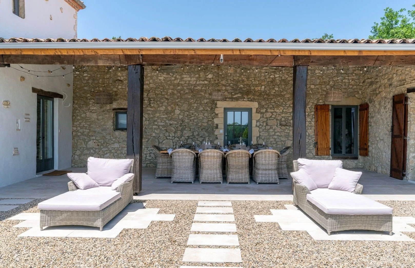 Outdoor patio with wicker lounge chairs and a dining table, stone walls with wooden shutters, and string lights under a tiled roof.