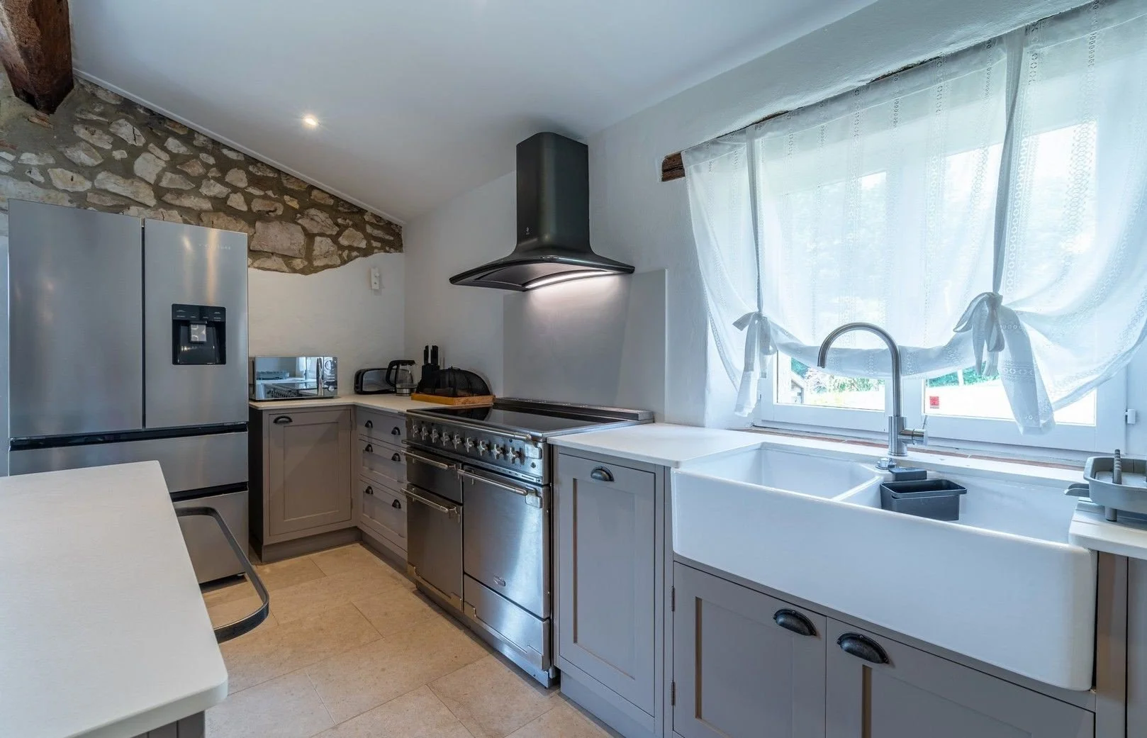 Kitchen with stone accent wall, stainless steel refrigerator, white farmhouse sink, window with sheer curtains, stove with hood, and countertop appliances.