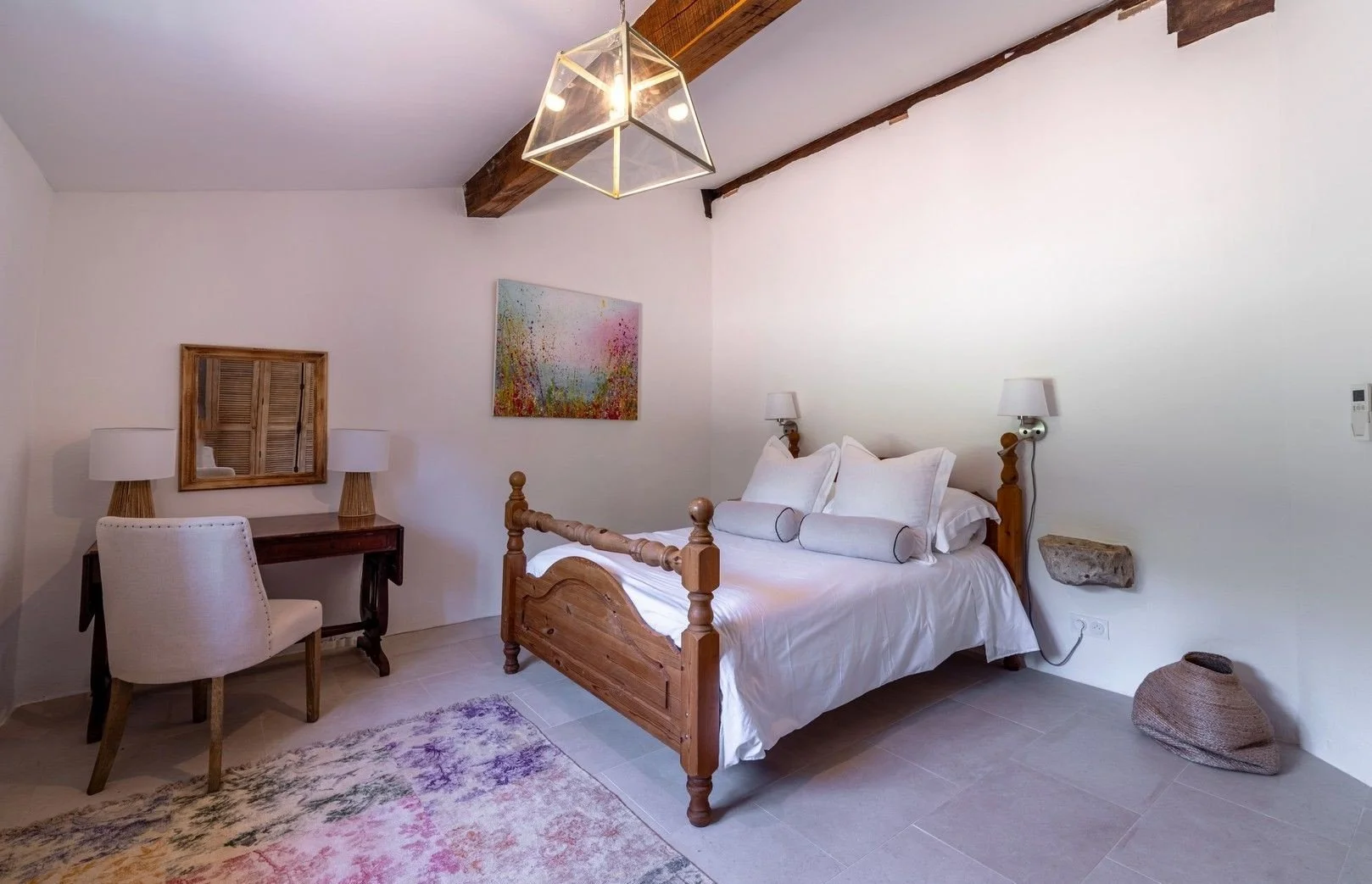 A bedroom with a wooden bed, white bedding, and multiple pillows. A wall-mounted lamp on each side of the bed, a colorful painting above the headboard. A white upholstered chair, a desk with two lamps, a mirror, a decorative basket on the floor, and 