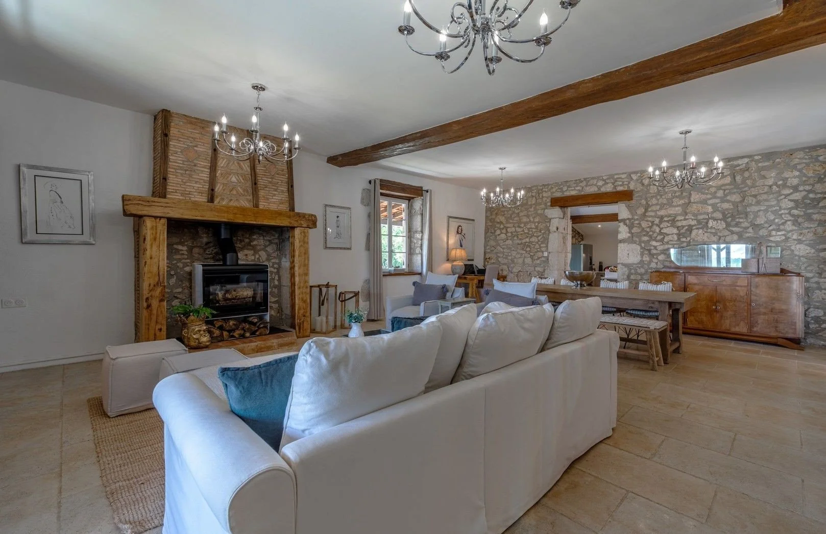 Living room with a stone fireplace, white sofa with blue and gray pillows, wooden accents, chandeliers, a window with curtains, and a dining area in the background.