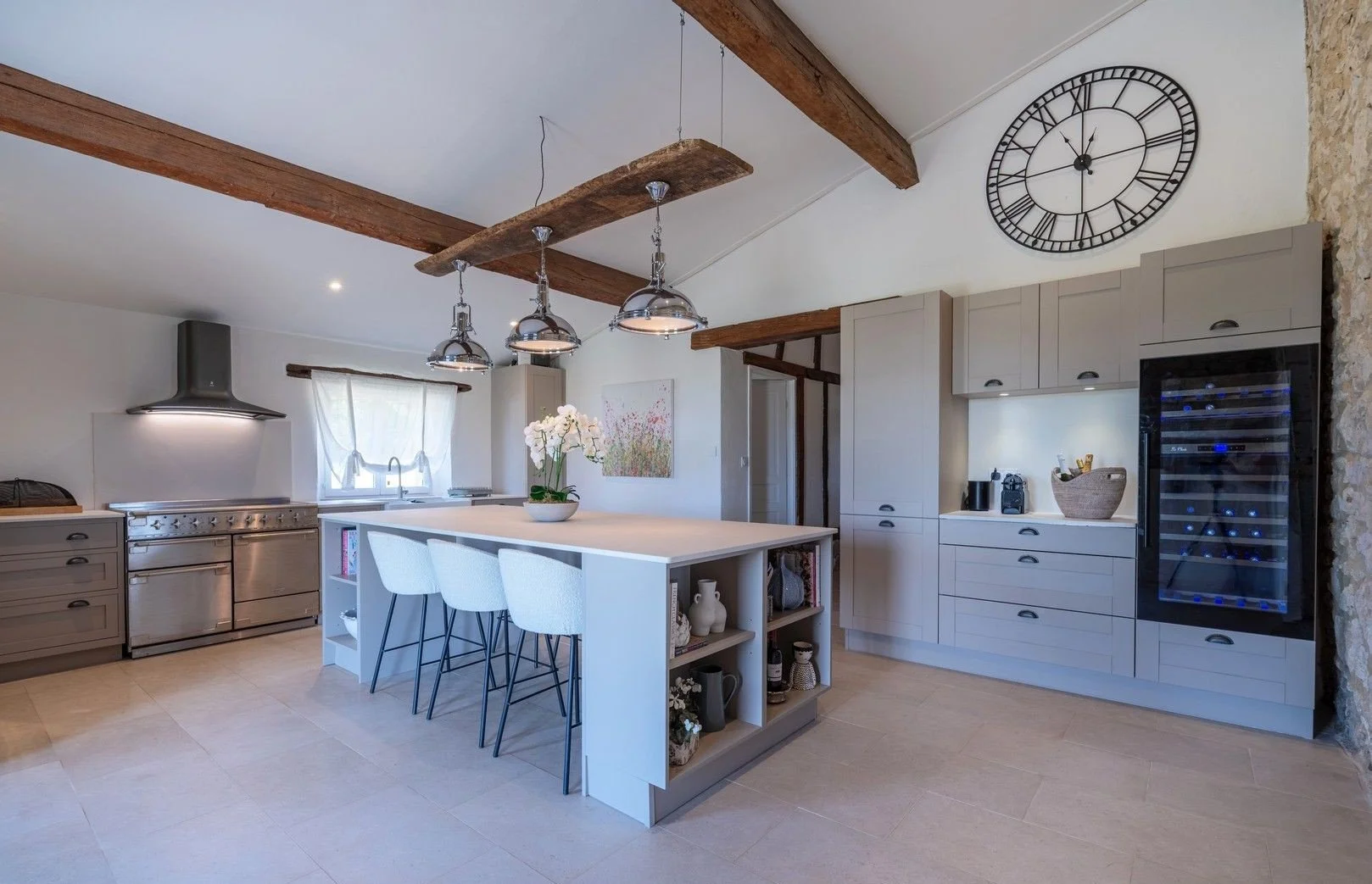 Modern kitchen with a white island, bar stools, stainless steel appliances, a large wall clock, wooden ceiling beams, and a wine cooler.