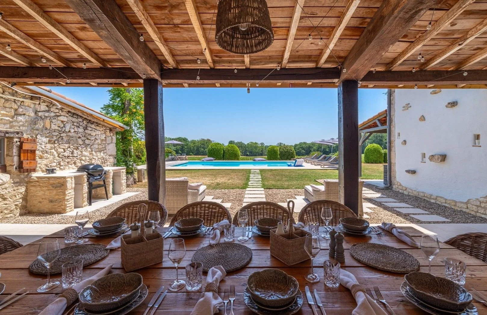 A dining table set with plates, wine glasses, and napkins, under a wooden patio ceiling, with an outdoor pool and lounge chairs in the background.