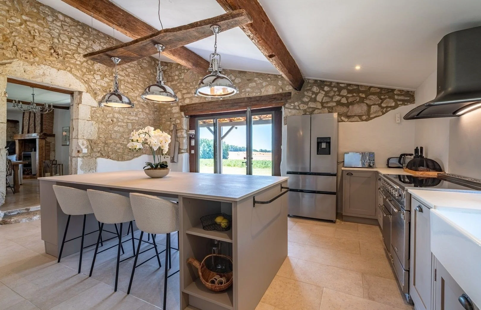 Kitchen with stone walls, wooden beams, kitchen island with chairs, stainless steel refrigerator, large wine fridge, oven, and a large window with a view of a green field.