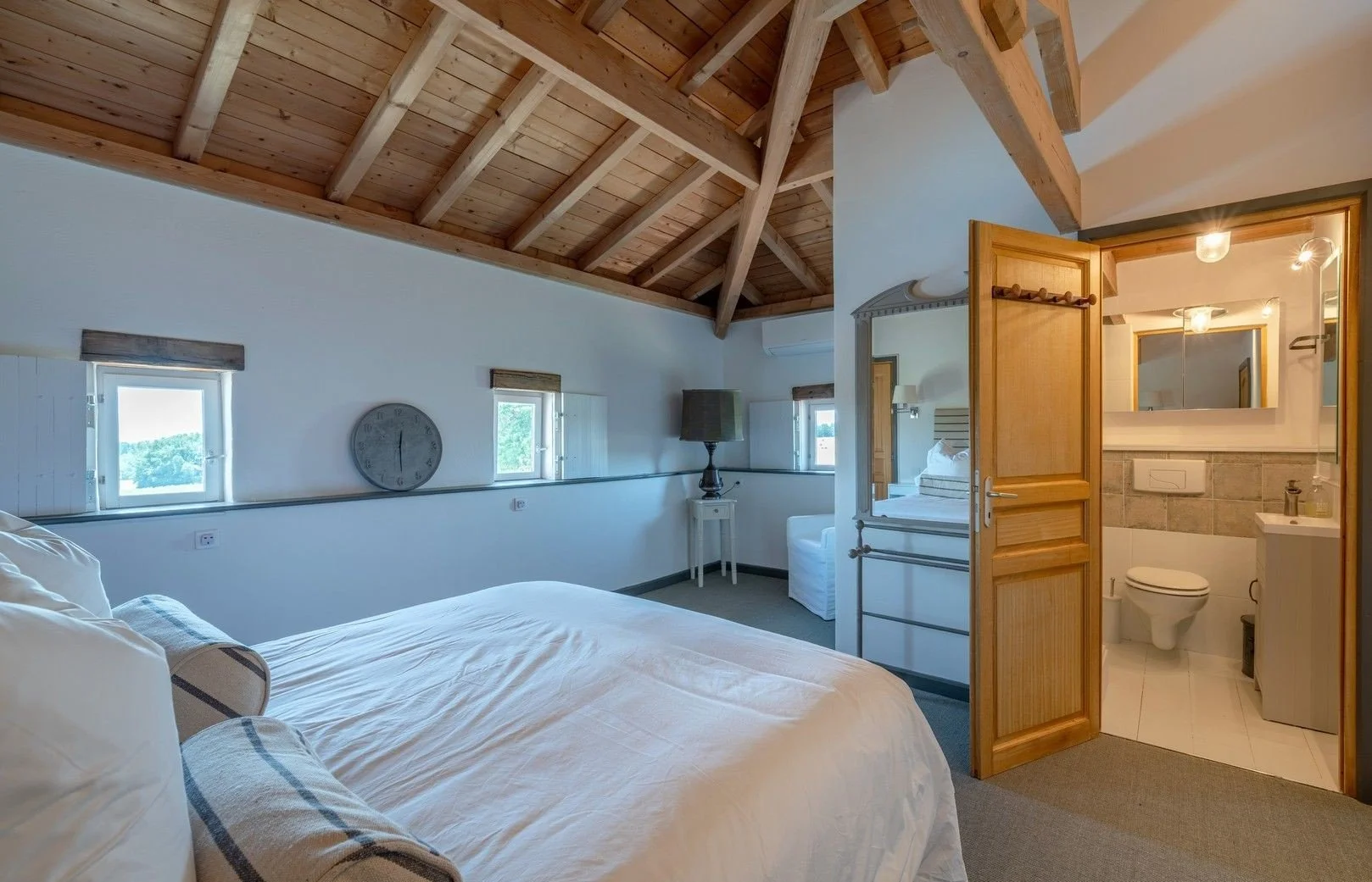 A cozy bedroom with a vaulted wooden ceiling, small windows, a bed with white linens, and a view of a bathroom through an open door.