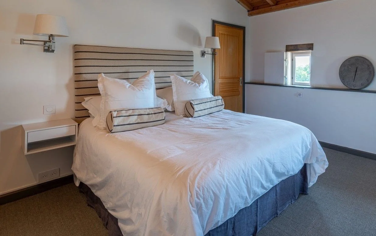 A neatly made bed with white bedding, decorative pillows, and a striped headboard in a cozy bedroom with wooden ceiling and door, small window, and a round wall clock.