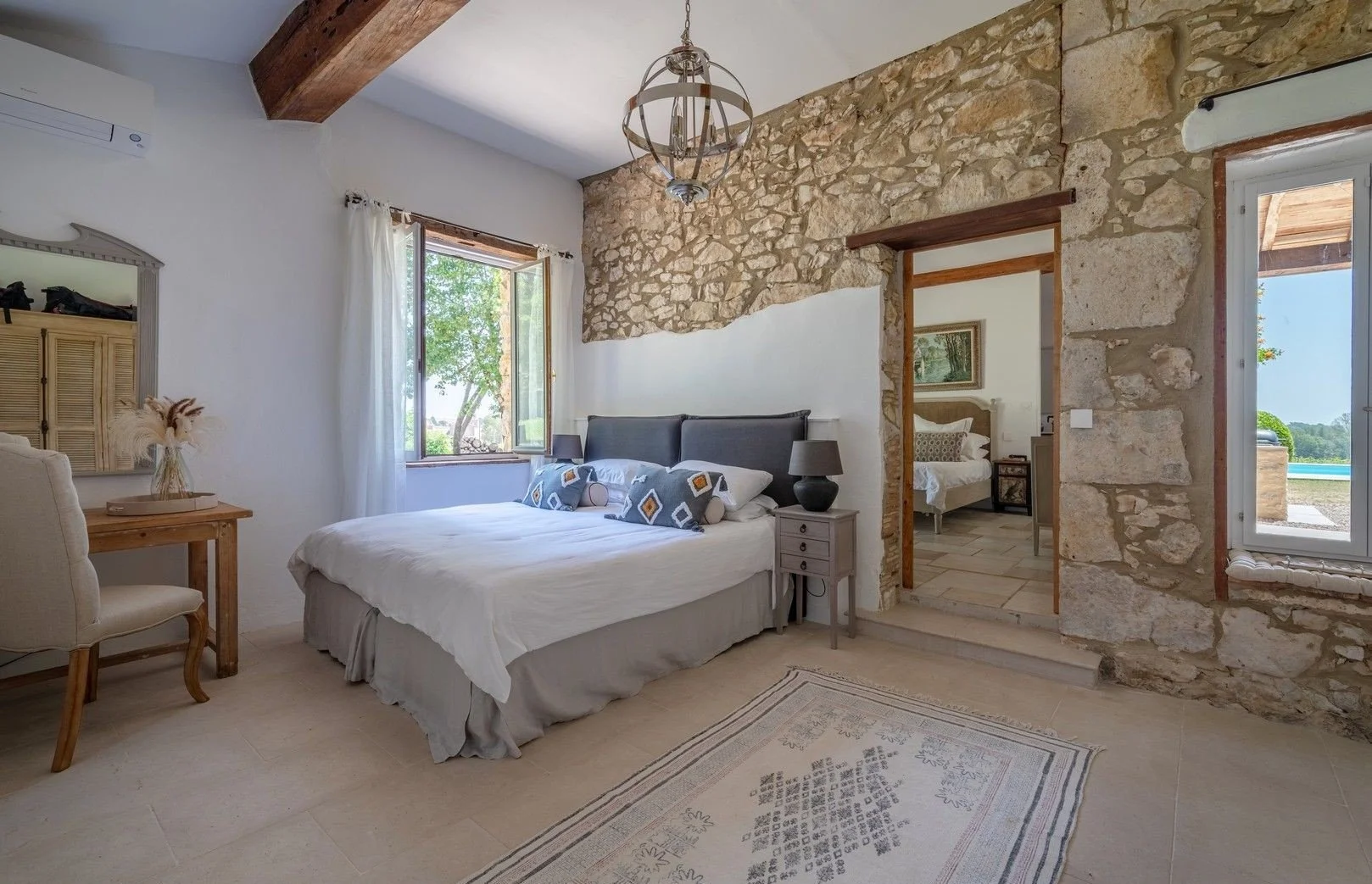 A bedroom with a stone and white wall, a bed with gray headboard and patterned pillows, a window with white curtains, a nightstand with black lamps, a wooden desk with a chair, and a partially visible doorway leading to another bedroom. There is an o