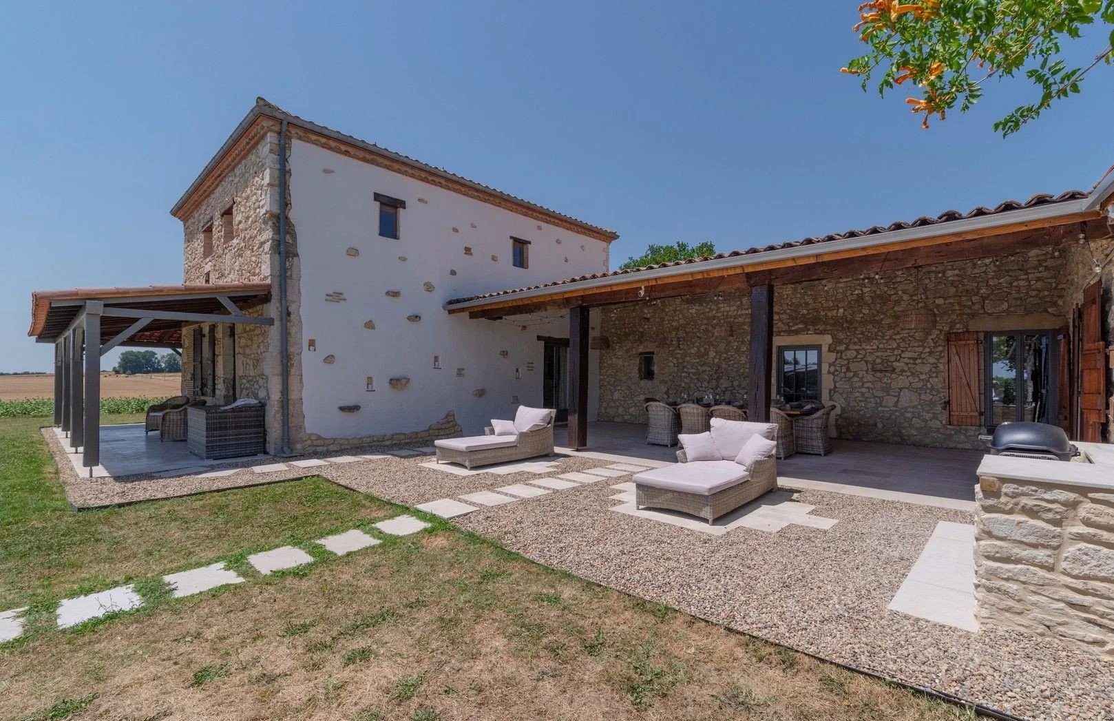 View of a rustic house with stone walls, a wooden roof, and an outdoor patio area furnished with cushioned outdoor furniture, set in a rural landscape under a clear blue sky.