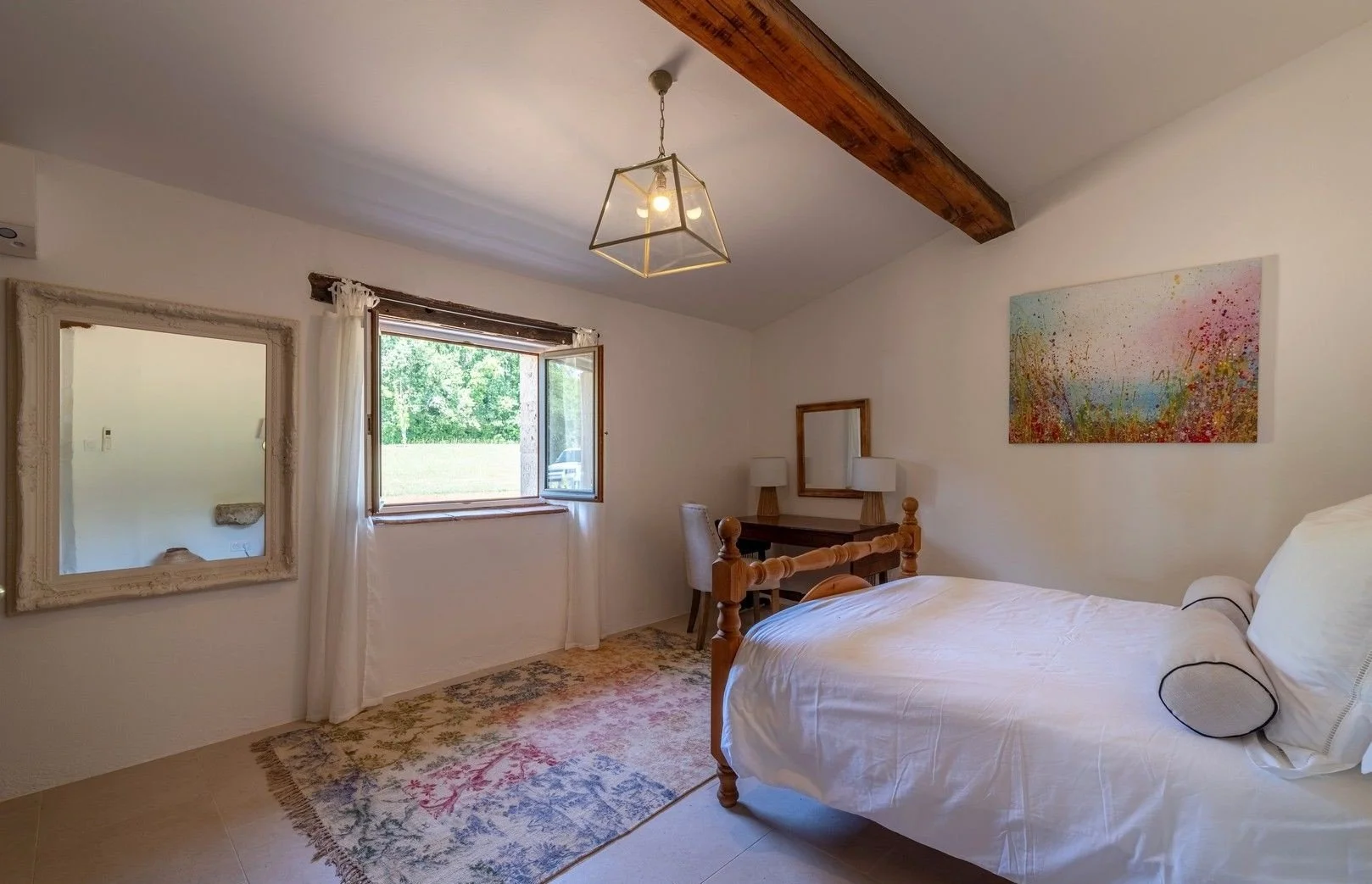 A cozy bedroom with a wooden bedframe, white bedding, and a decorative pillow. There is a colorful abstract painting on the wall, a window with white curtains, a vintage-style mirror, a wooden desk with two lamps and a mirror, a patterned rug, and a 