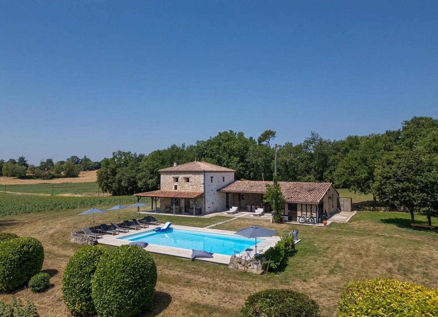 A rural property with stone buildings, a swimming pool with lounge chairs and umbrellas, on a sunny day with green trees and fields in the background.