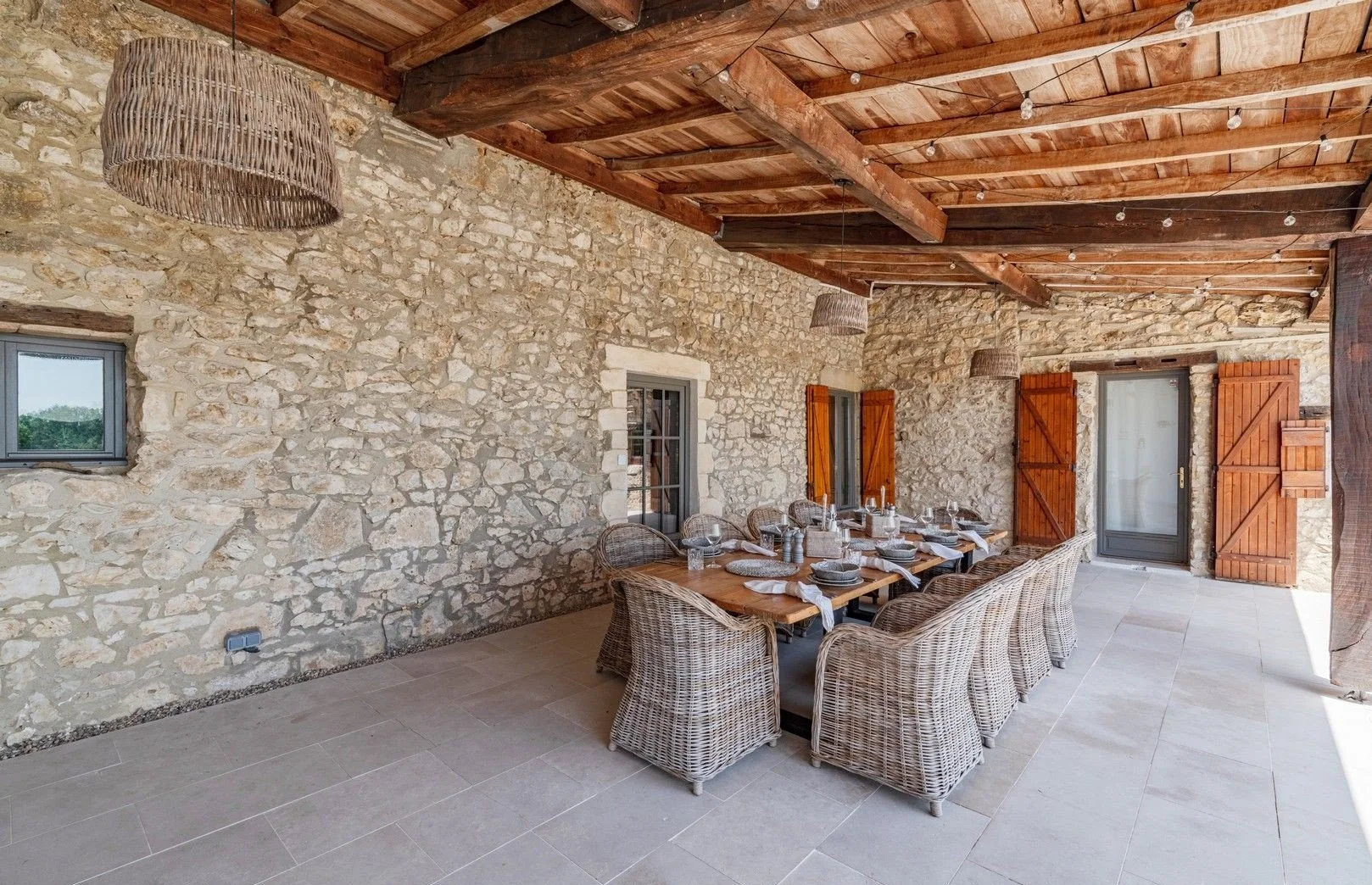 Rustic outdoor dining area with a long wooden table, wicker chairs, stone walls, wooden ceiling, and open wooden shutters.