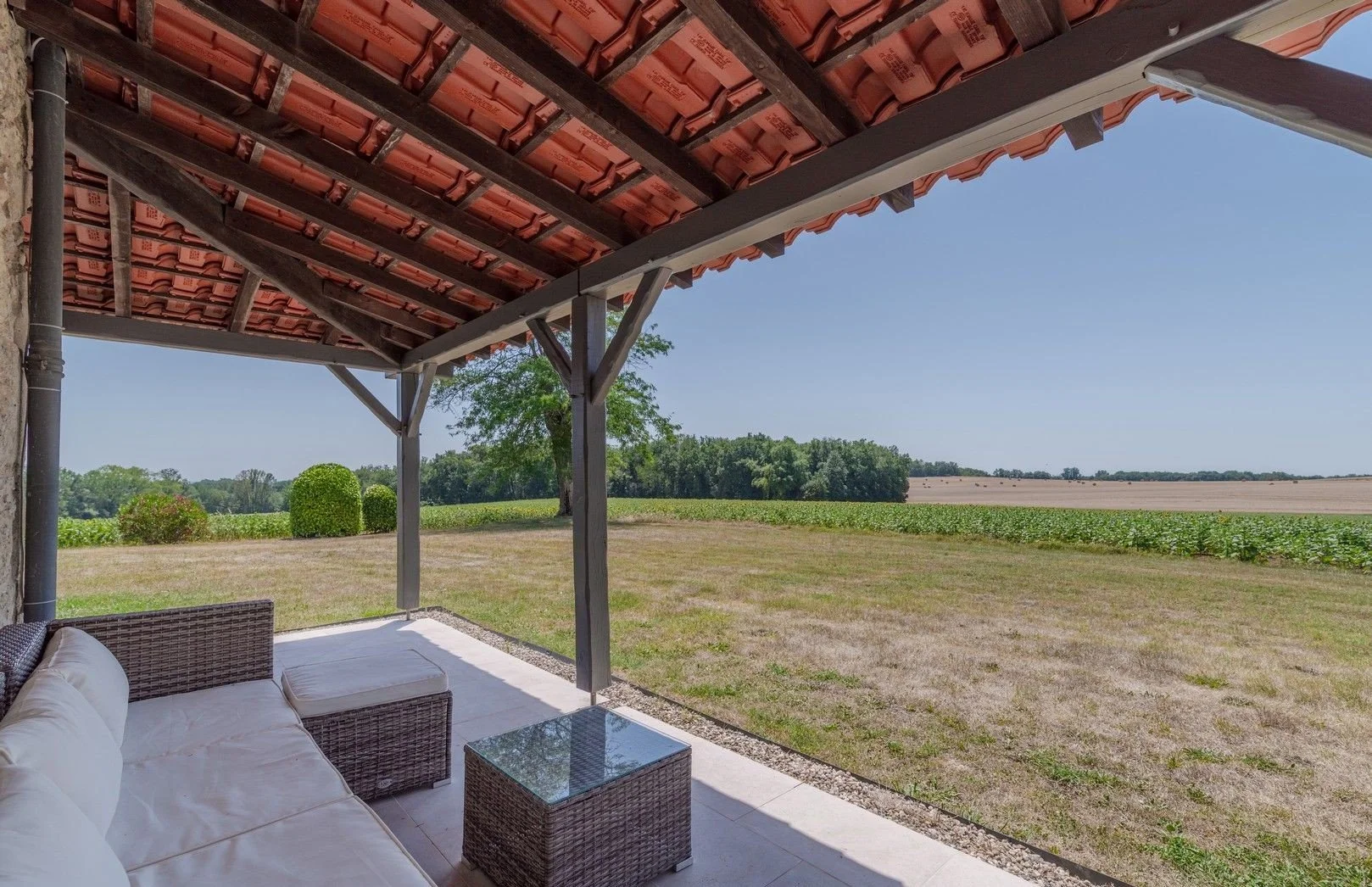 A covered outdoor patio with wicker furniture and a glass-topped table, overlooking a spacious field with trees in the distance under a clear blue sky.
