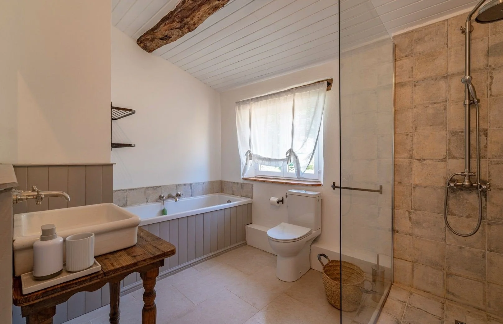 A bathroom with a rustic wooden beam on a white-paneled ceiling. It features a bathtub with a faucet and a small window with white curtains, a toilet with a toilet paper holder, a glass-enclosed shower with beige tiled walls, a wicker laundry basket,