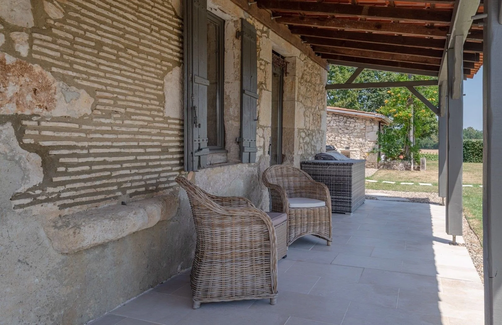 Photo of a rustic outdoor porch with wicker chairs, stone and brick wall, and a scenic view of a grassy yard with trees in the background.