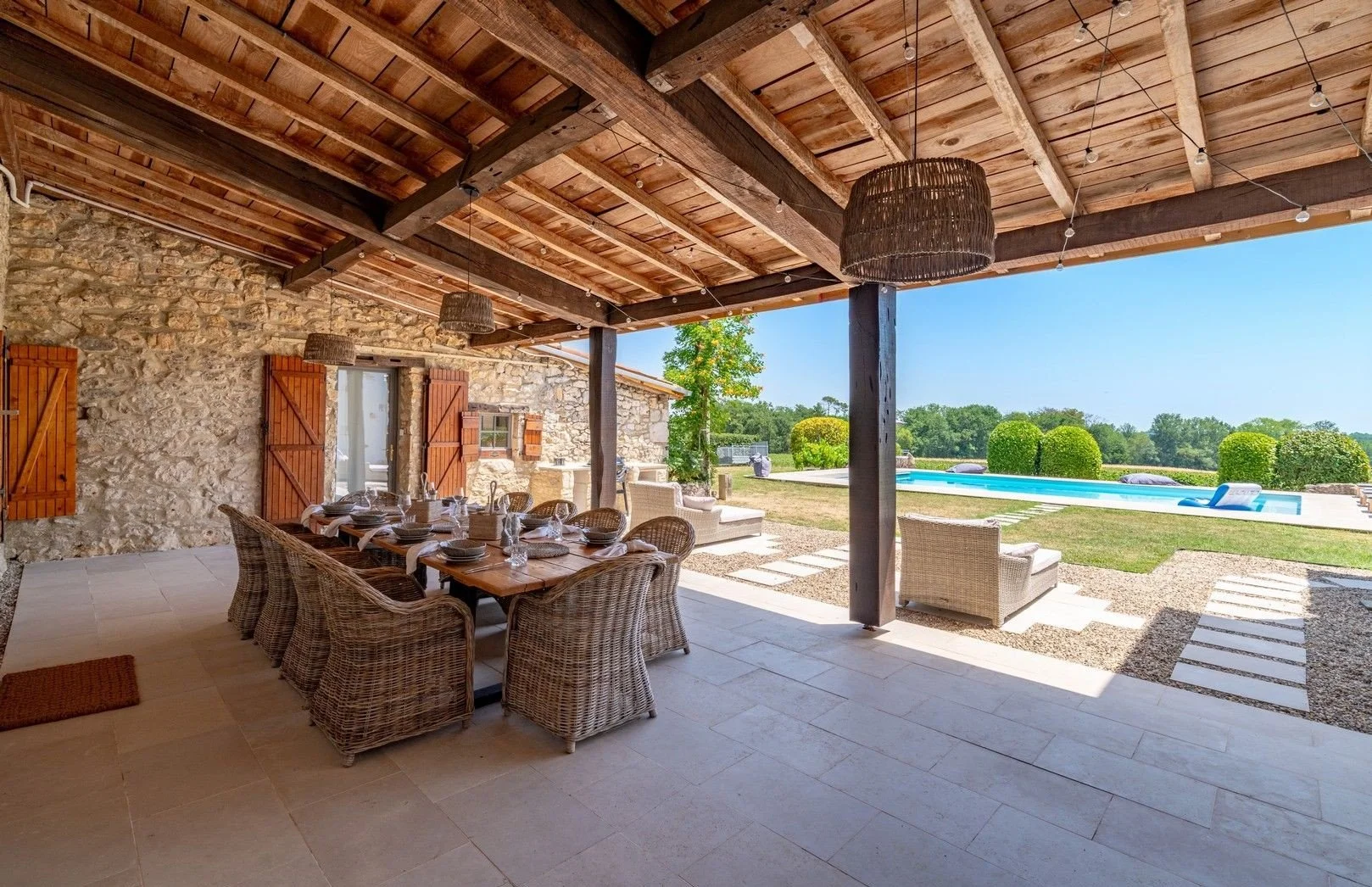 Outdoor patio with a dining table set for a meal, wicker chairs, and a stone wall. Beyond the patio, there is a swimming pool, green shrubs, and a grassy area under a clear blue sky.