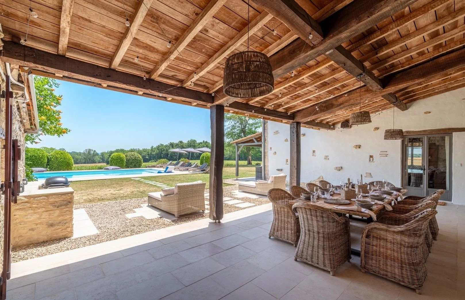 Outdoor patio area with dining table and chairs, overlooking a backyard with a swimming pool, lounge chairs, and umbrellas on a sunny day.
