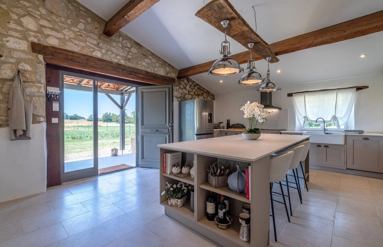 A spacious kitchen with a large white island, a stainless steel refrigerator, and stone walls with wooden beams. Open sliding doors lead to a patio overlooking fields. There are curtains on a window above the sink and modern pendant lighting.