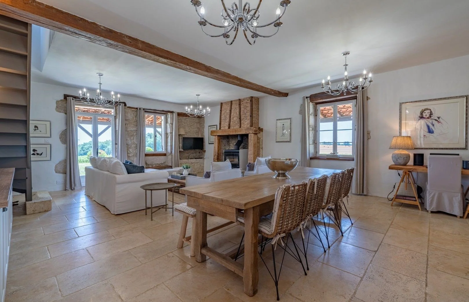 Living and dining room with stone fireplace, chandelier lighting, white sofas, wooden dining table with woven chairs, framed artwork, and large windows with views of outdoors.
