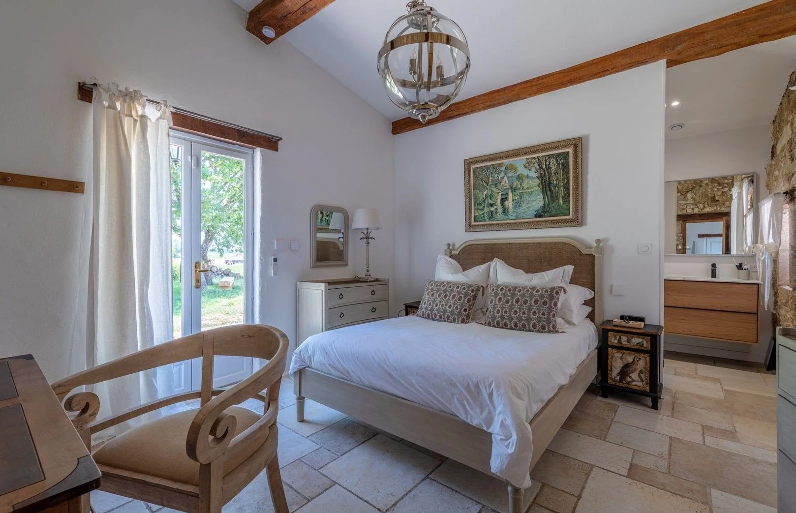 A cozy bedroom with a large bed featuring white bedding and patterned pillows, a wooden headboard, a white dresser with a mirror and a lamp, a small dark side table, a window, and a glass-paneled door with curtains leading outside. The room has rusti