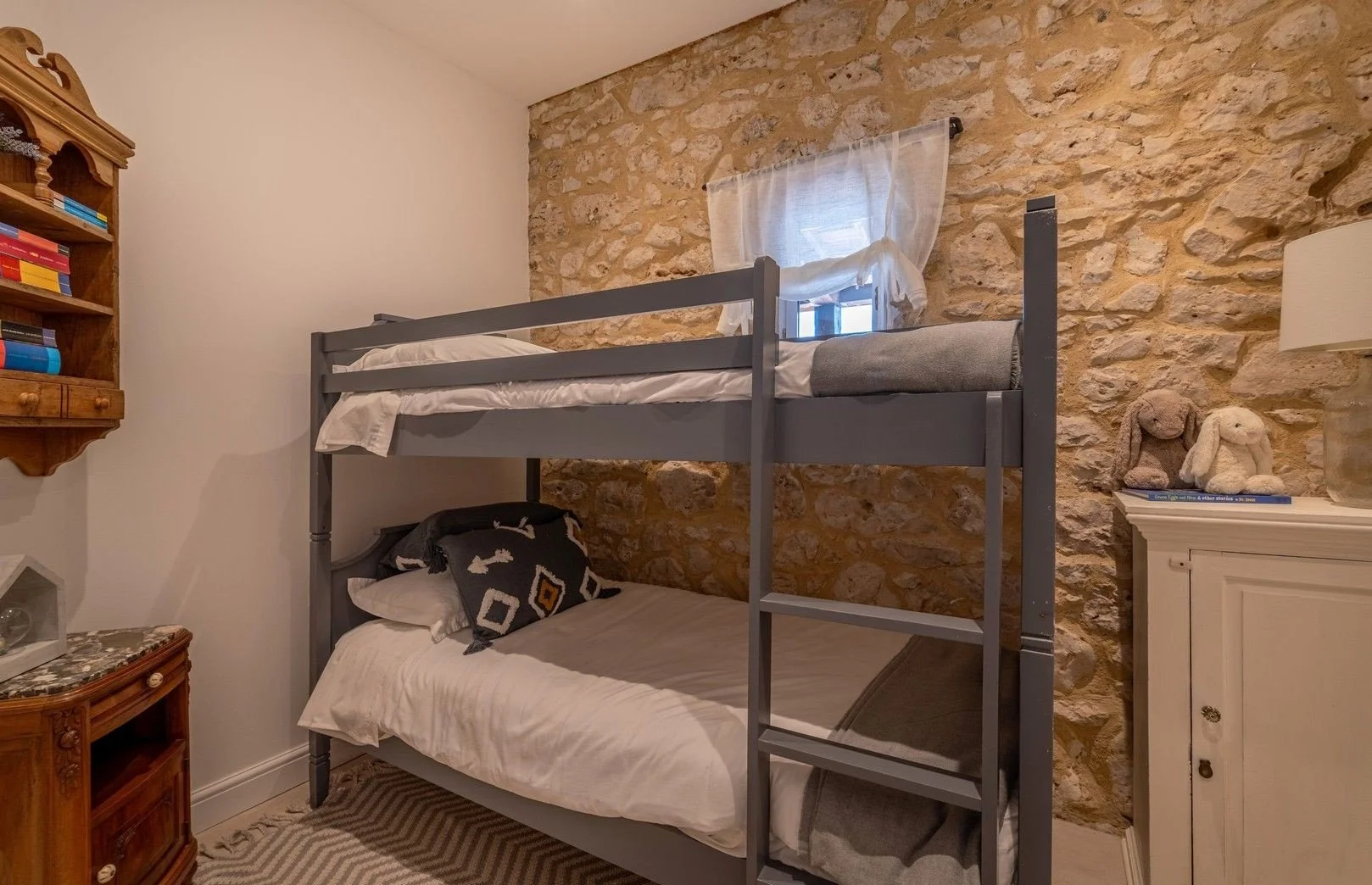 A bedroom with a stone accent wall, white painted wall, wooden bookshelf, and white furniture. Contains a bunk bed, with a lower bed with a black pillow and an upper bed with plain bedding. A small window with sheer curtain is above the bunk bed. The