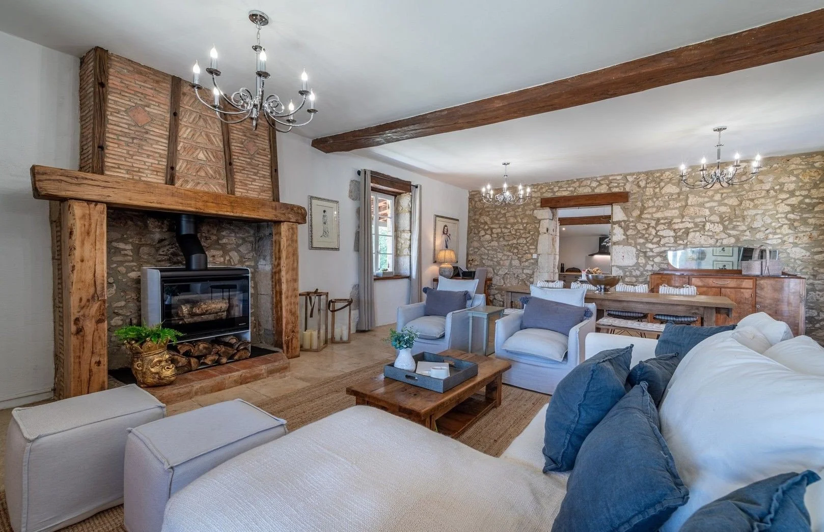 Living room with stone walls, wooden beams, a fireplace with logs, white sofas with blue and white cushions, a wooden coffee table, chandeliers, and large windows with curtains.