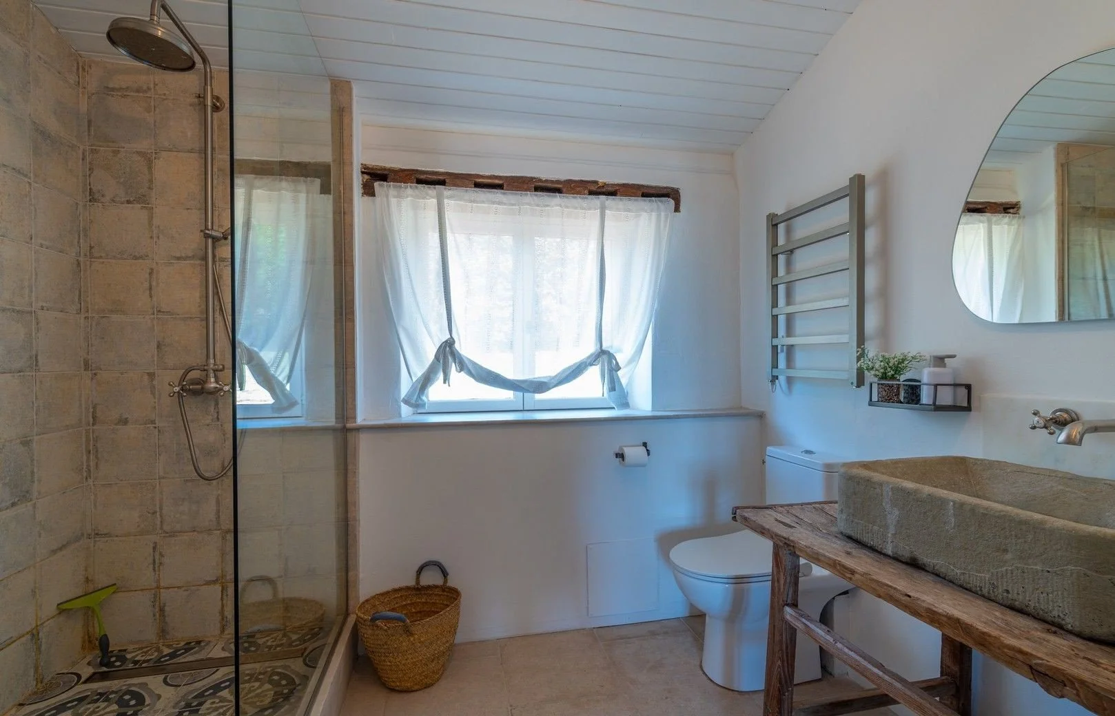 A rustic bathroom with a walk-in shower on the left, a window with white curtains in the center, and a toilet, sink, and mirror on the right. The decor features natural wood and stone elements.
