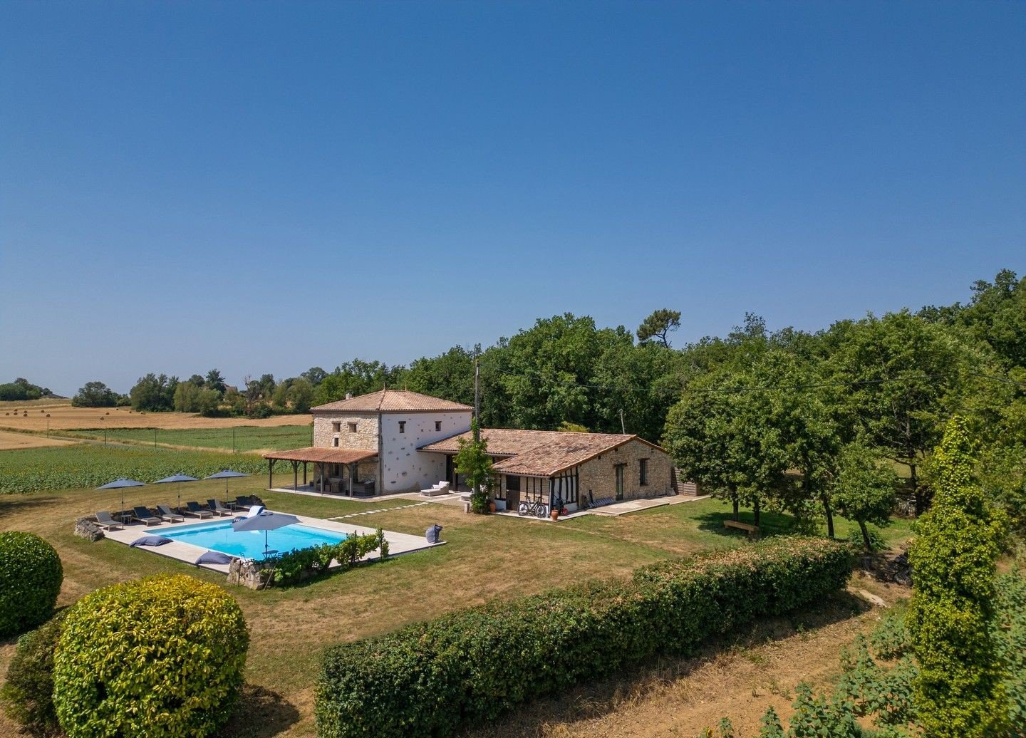 A rural house with a swimming pool, surrounded by greenery, trees, and farmland under a clear blue sky.