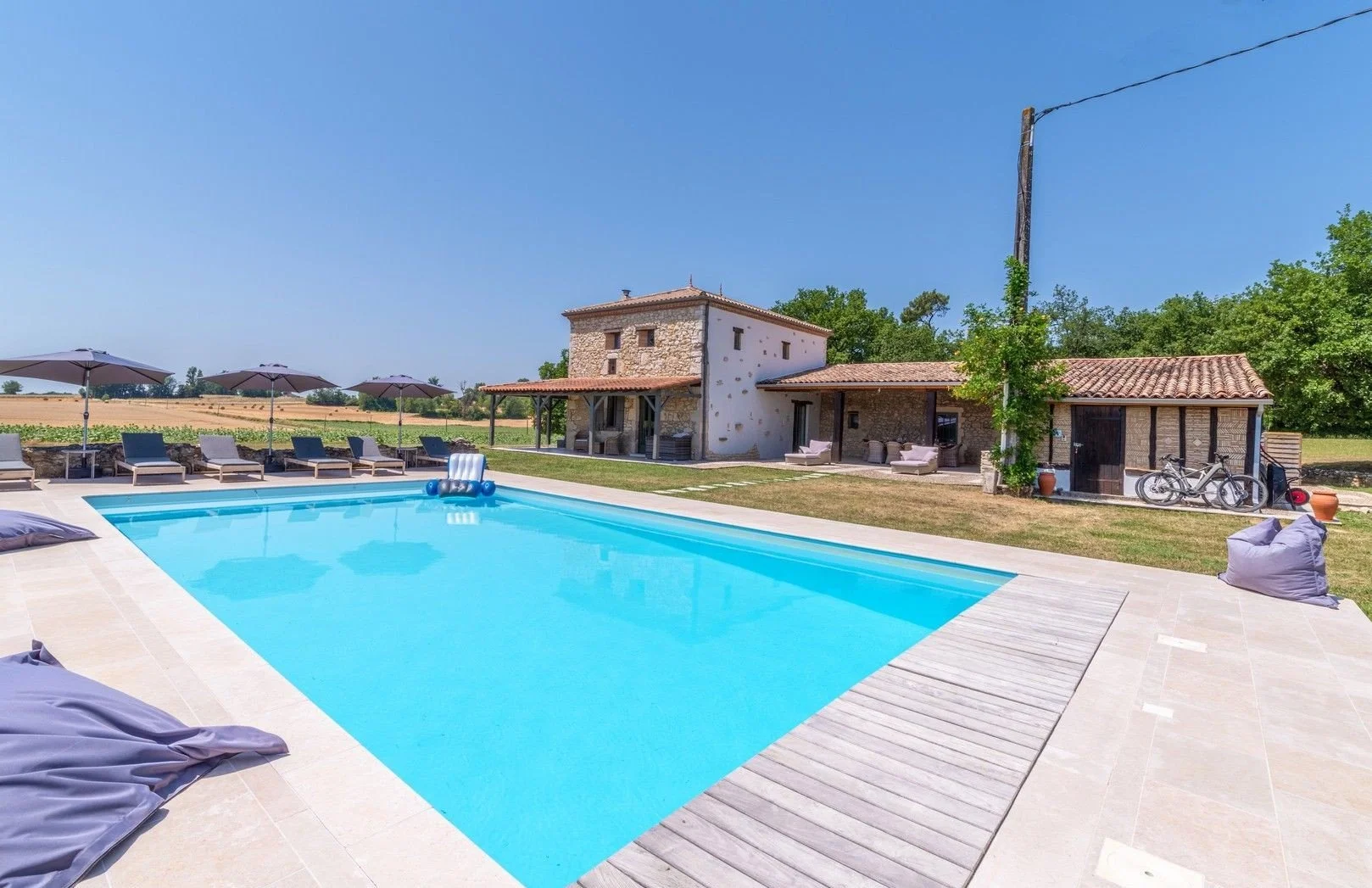 A backyard with a swimming pool, lounge chairs with umbrellas, bean bags, a rustic stone house, and a surrounding rural landscape with fields and trees under a clear blue sky.