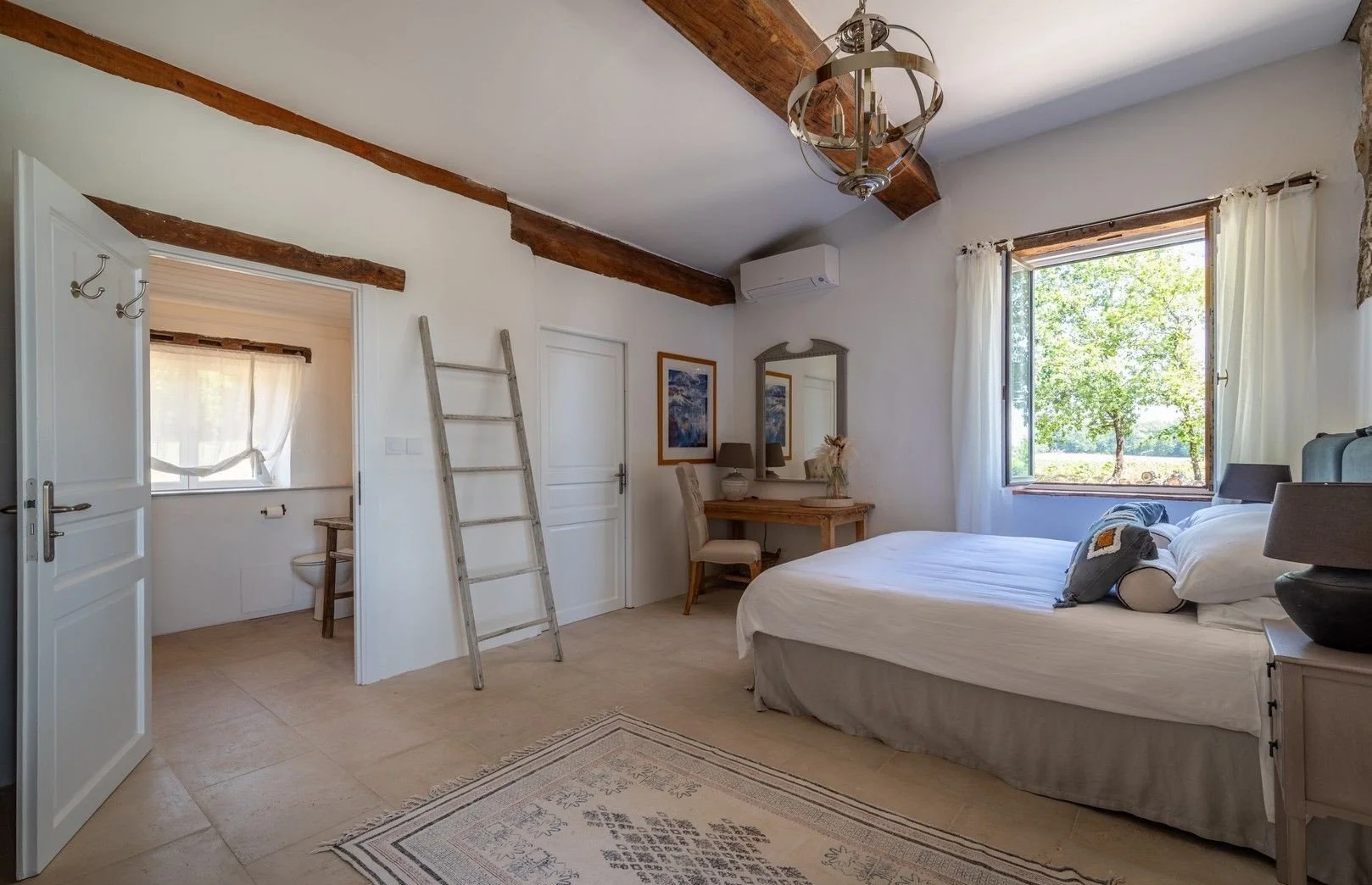 A bedroom with a large bed near a window showing a tree outside, a wooden desk with a mirror, framed pictures, a chair, a ladder, and a small bathroom area with a toilet visible through an open door.