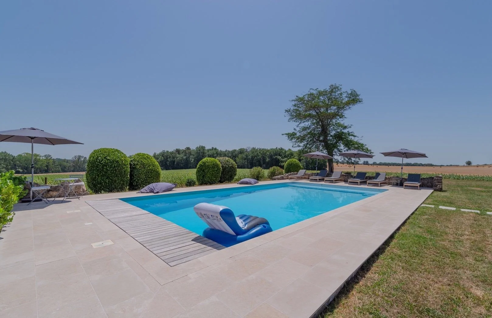 Outdoor scene with a swimming pool, lounge chairs, umbrellas, manicured bushes, and open fields under a clear blue sky.