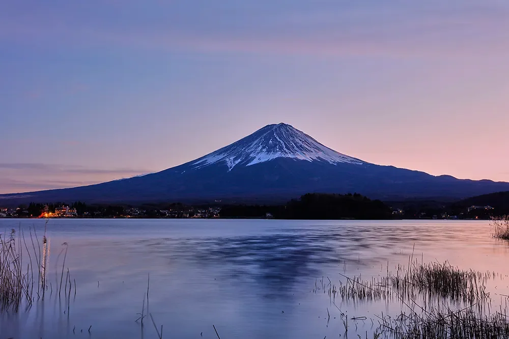 Mount Fuji at magic hour seen across Lake Kawaguchiko in Japan, with a snow-capped summit, still water, shoreline reeds, and village lights under a violet-pink sky.